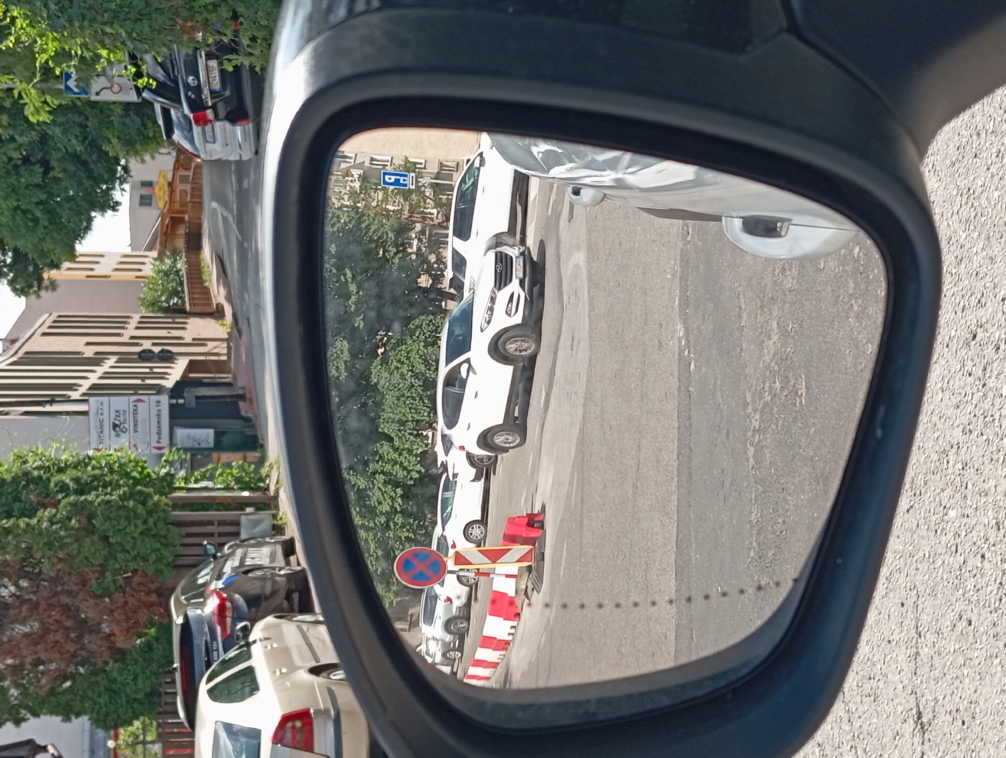 The image is a reflection in a car's side mirror, showing a street scene in Nitra, Slovakia. A white SUV is parked on the right side of the road, while a white sedan is parked on the left. A temporary barrier with red and white markers is set up in the middle of the road, indicating a construction or road closure. A no-parking sign is visible above the barrier. In the background, buildings and trees line the street. The overall environment appears to be a residential or commercial area on a sunny day.
