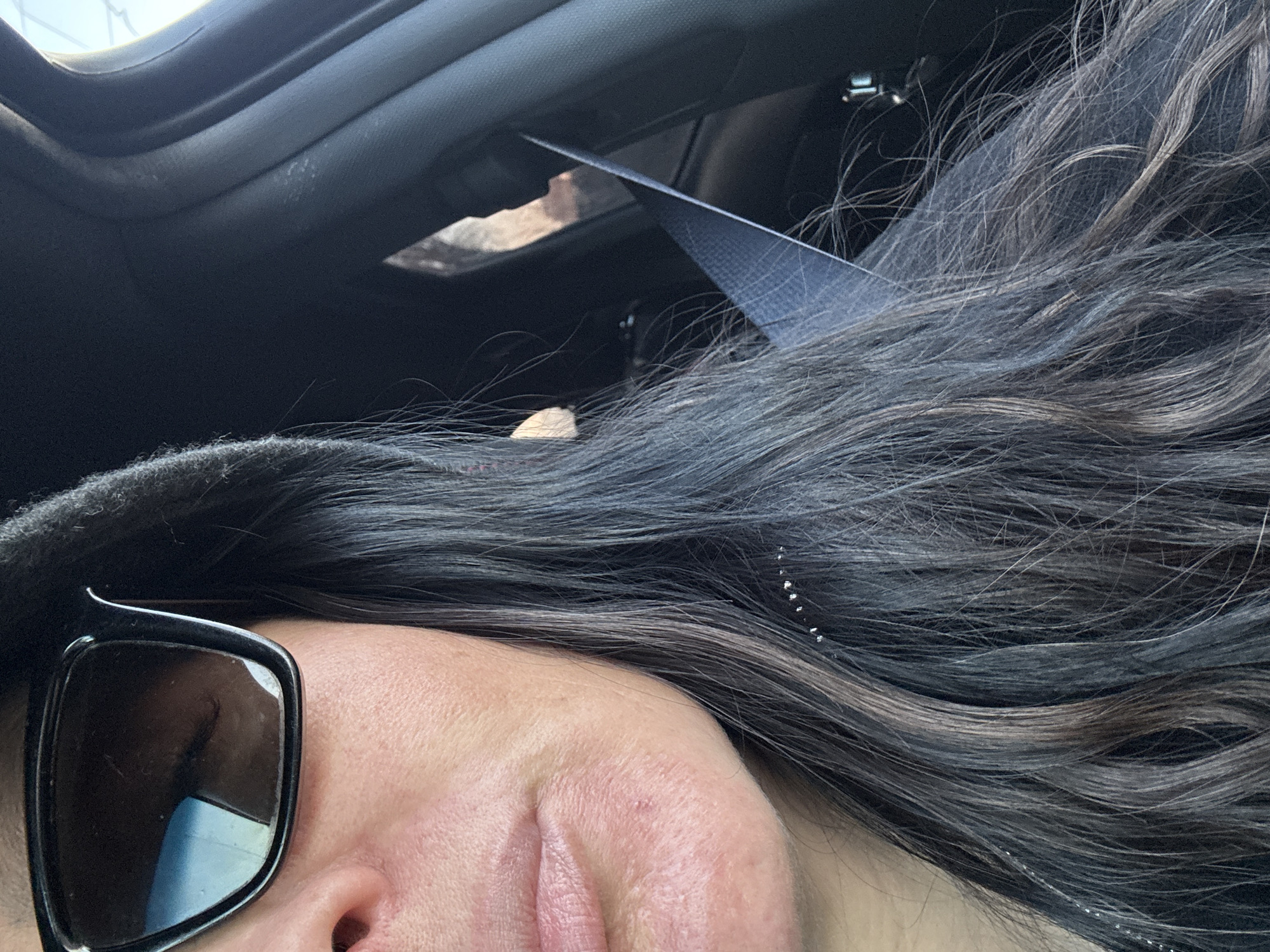 A close-up shot, seemingly taken from inside a car, features a person wearing sunglasses and a hat. The person's face, including their eye reflected in the sunglasses, cheek, nose, and lips, occupies the lower-left portion of the frame. Wavy, dark hair with lighter highlights cascades across the center and right. A car seatbelt cuts across the top of the hair. The car's interior, including the roof and sunroof, is visible in the background. The setting appears to be inside a vehicle. There is insufficient information to determine the location as Chaikovsky, Russia.