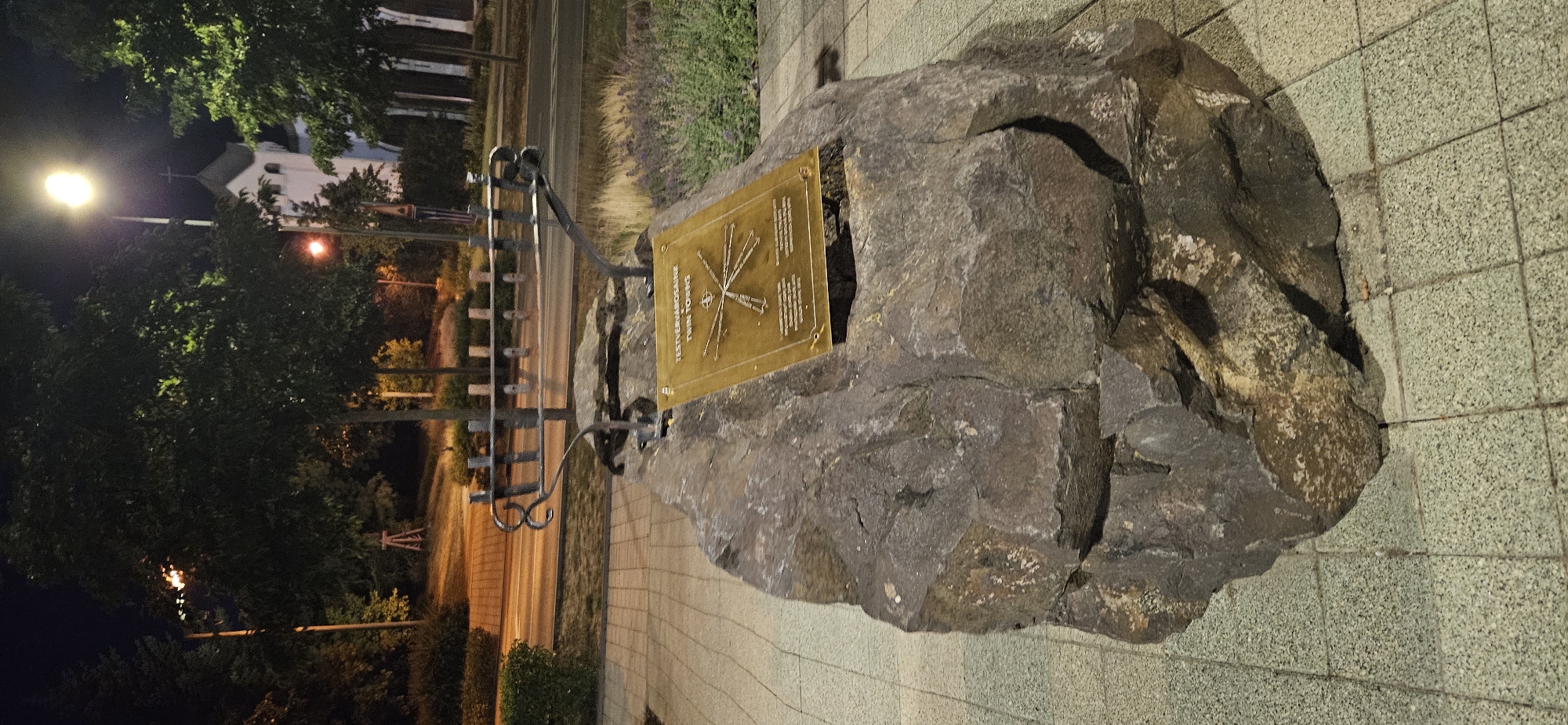 The image shows a large, rough-textured rock with a bronze plaque affixed to it. The plaque is rectangular and appears to have text and a diagram etched onto its surface. Above the rock, a metal sculpture resembling a menorah or a coat rack is attached to a base that is integrated with the rock. In the background, there are trees, a street with a car, and a building with a steeple and a cross, suggesting a public outdoor space, possibly in a town or city. The lighting indicates it is either dusk or nighttime.

The scene is located in Hévíz, Hungary. Specifically, this is the "Twin Towns" memorial located on the corner of Petőfi utca and Fodor József utca, near the town hall and the Roman Catholic Church. The plaque commemorates Hévíz's twin towns. The diagram on the plaque depicts rays emanating from a central point, with names and possibly distances to different towns. The metal sculpture above the rock is a symbolic representation of connections between towns. The overall presentation suggests a monument or public art installation.