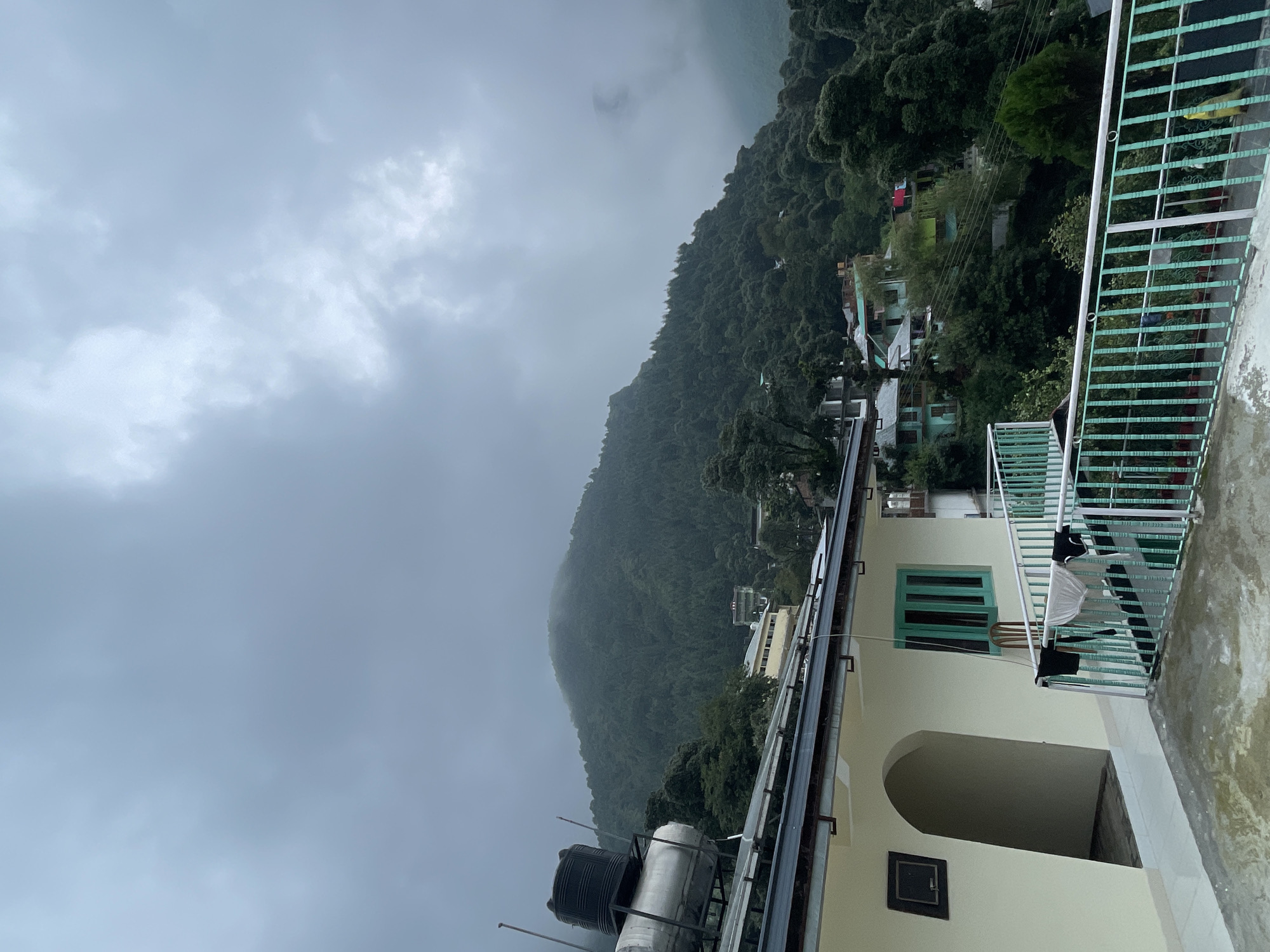 The image depicts a view from a rooftop in Bhagsu, India, overlooking a hillside covered in lush green trees. The sky is overcast with heavy gray clouds, suggesting a possibly damp or cool day. In the foreground, a section of a building with a pale yellow exterior and an arched doorway is visible. A balcony railing, painted in a teal and white striped pattern, runs along the front of the building. Laundry, including dark and light colored garments, hangs on a line supported by the railing. A water tank and a solar water heater are mounted on the roof of the building to the left. Further down the hillside, several houses with colorful roofs are nestled amongst the trees. The distant mountains are shrouded in mist and clouds, creating a dramatic, atmospheric backdrop.