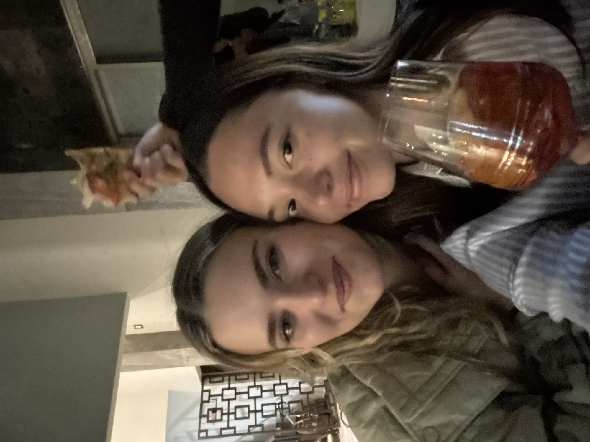 The image captures two females in a dimly lit indoor environment. The female on the left has long, wavy light-colored hair and is smiling, wearing a light olive-green quilted jacket. The female on the right has long, dark hair and is also smiling, wearing a striped shirt. She holds a stemmed glass containing a reddish-orange beverage with ice and citrus slices in her right hand. Above and behind the right female's head, the hand of an unseen third person holds a partially eaten slice of pizza. The background on the left features a dark, geometrically patterned screen or room divider, with light-colored walls beyond. The background on the right is darker, showing faint lights and obscured objects, suggesting a bar or dining area. The specific location within the city of Puebla, Mexico, cannot be determined from the visual evidence. Both visible subjects are looking towards the camera, posing for a close-up photograph.