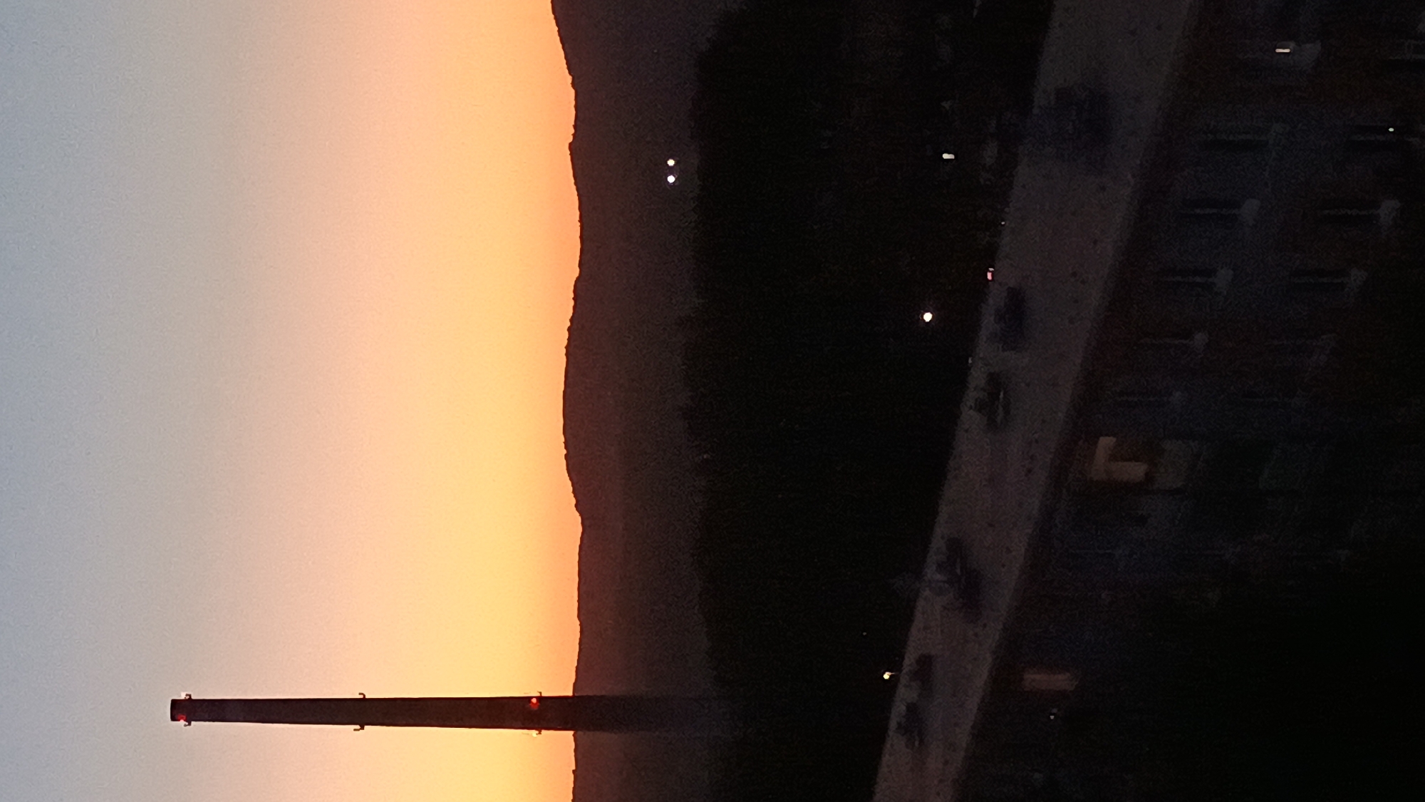 An elevated view of a cityscape at dusk shows a tall industrial smokestack on the left, silhouetted against a gradient sky transitioning from pale blue at the top to orange near the horizon. Below the smokestack and extending across the frame is a dark, hilly landscape covered in trees. In the foreground, a long, flat-roofed building with visible windows, some illuminated from within, stretches from the bottom left corner towards the right. Several faint lights are visible in the middle ground, two of which are clustered together. The overall impression is one of an industrial area at the edge of a town, observed from a residential building.