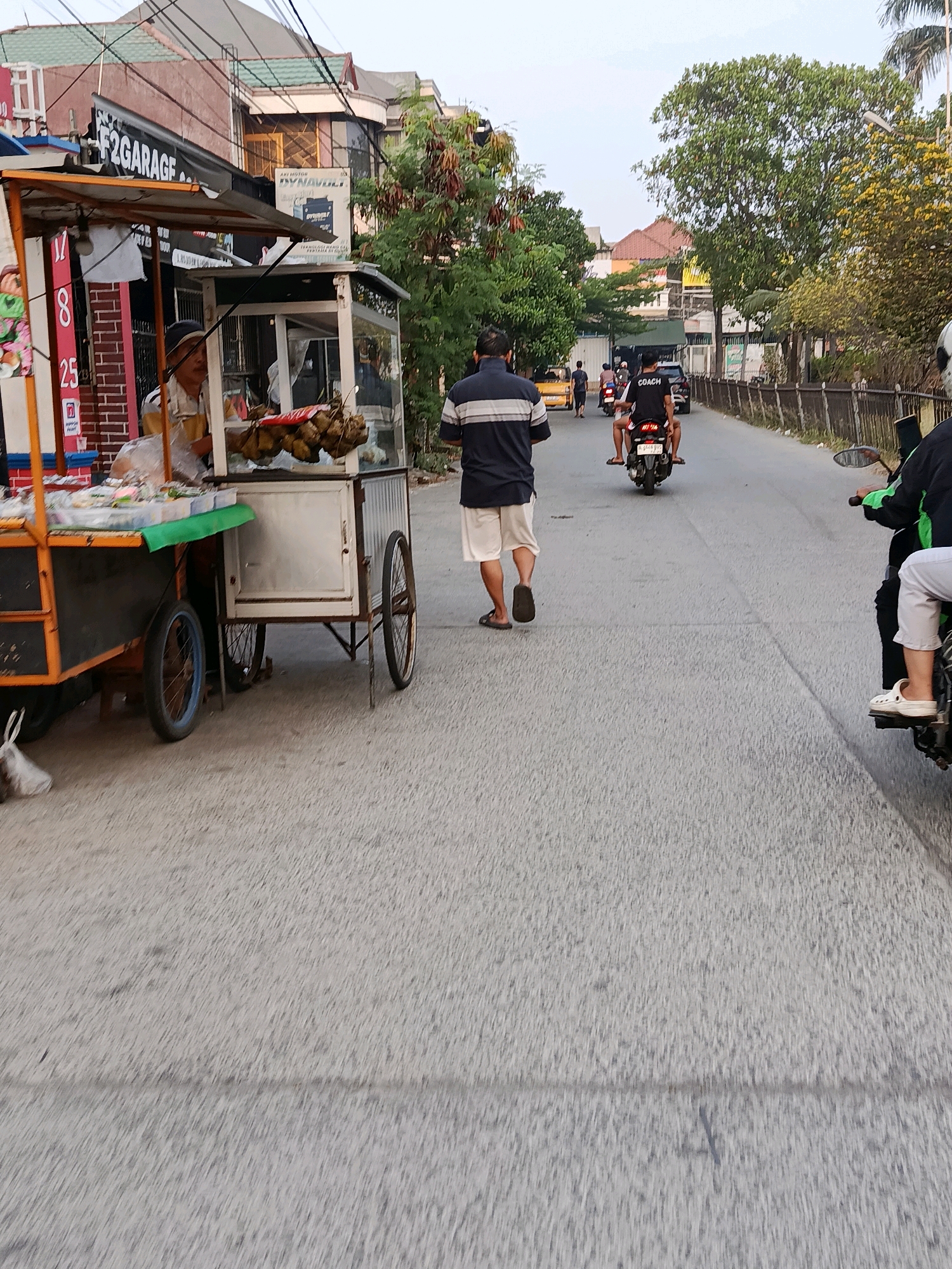 The image depicts a street scene in the city of Bekasi, Indonesia, likely during daylight hours as indicated by the lighting. In the foreground left, a food cart is visible, with a vendor attending to it. The cart is laden with food items and appears to be mobile, resting on wheels. Further down the street, several individuals are present. One man walks away from the camera, wearing a dark shirt with white stripes and light-colored shorts. Behind him, two people ride on a motorcycle, with the passenger wearing a black t-shirt that reads "COACH". Other motorcycles and vehicles are present on the road, suggesting a typical urban environment with traffic. Buildings line the street, some with signs, including one for "F2 GARAGE". Trees and vegetation are scattered along the roadside, indicating a mix of urban development and greenery. The overall impression is a snapshot of everyday life on a street in Bekasi.