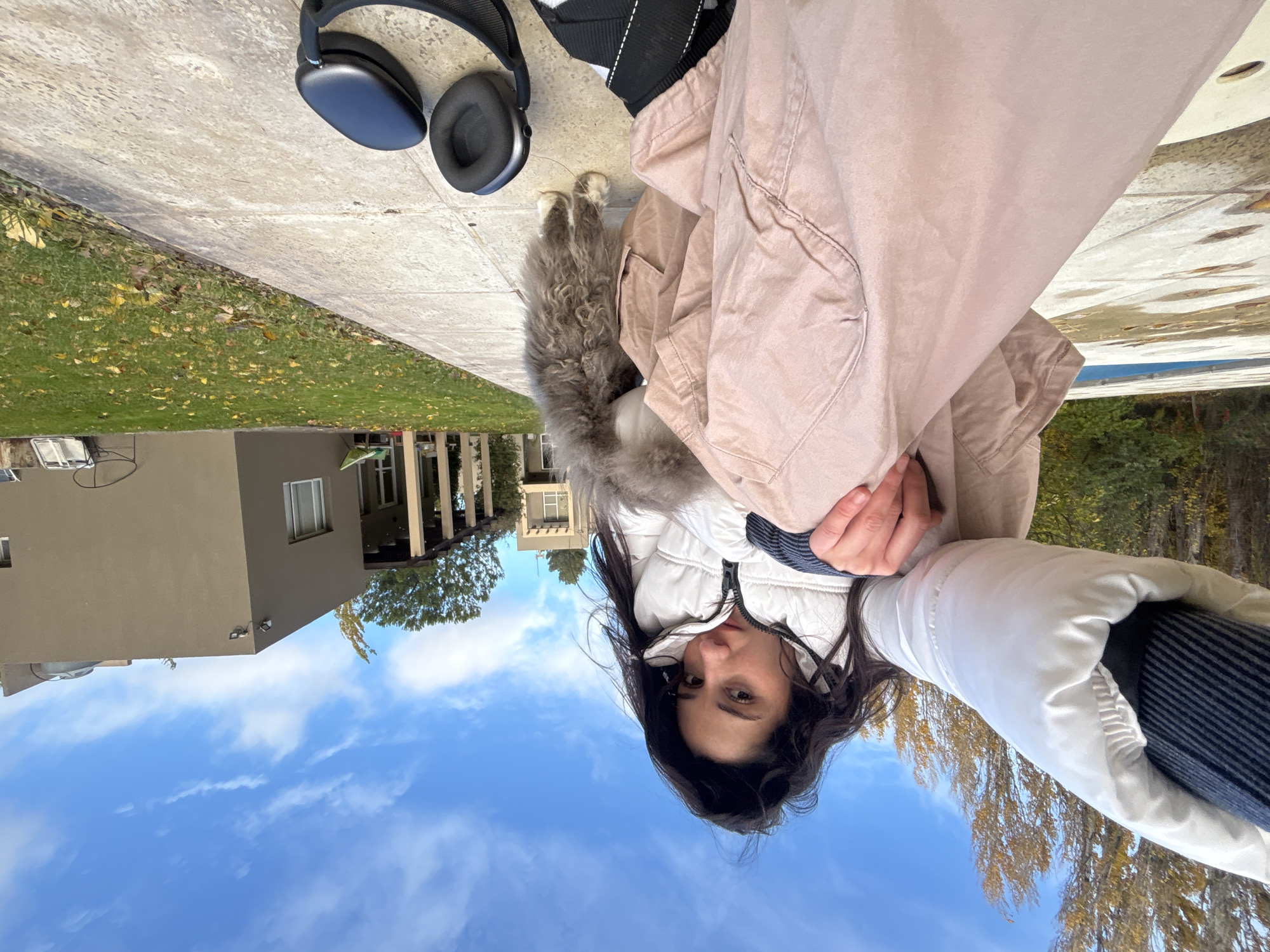 The image shows a person in a white and beige winter jacket with a fur hood, with a pair of black headphones nearby on a concrete surface. A fluffy, gray dog is standing between the person and the headphones. To the left, there is a green lawn and a building with windows. In the background, the sky is blue with some white clouds. The photo was taken in Trelew, Argentina.