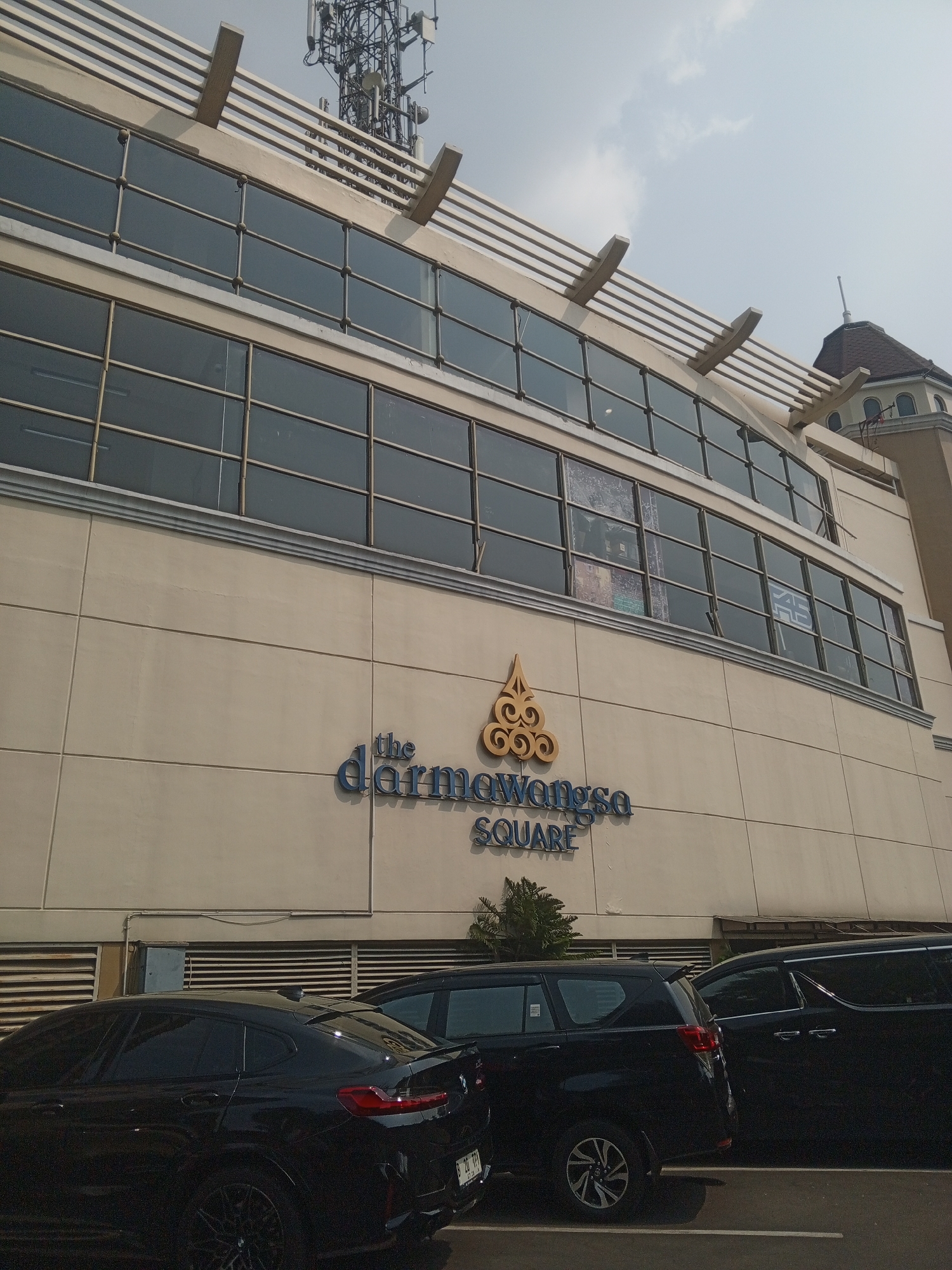 The exterior of a building is shown, with a prominent sign reading "the darmawangsa SQUARE" in blue lettering. The building has large, multi-paned windows on its upper floors, with a beige facade. A telecommunications tower is visible on the roof of the building. In the foreground, several dark-colored cars are parked. The sky above is hazy with some clouds. The scene appears to be a street view in a city. The building is likely located in South Jakarta, Indonesia, given the sign's name and the architectural style often seen in that region.