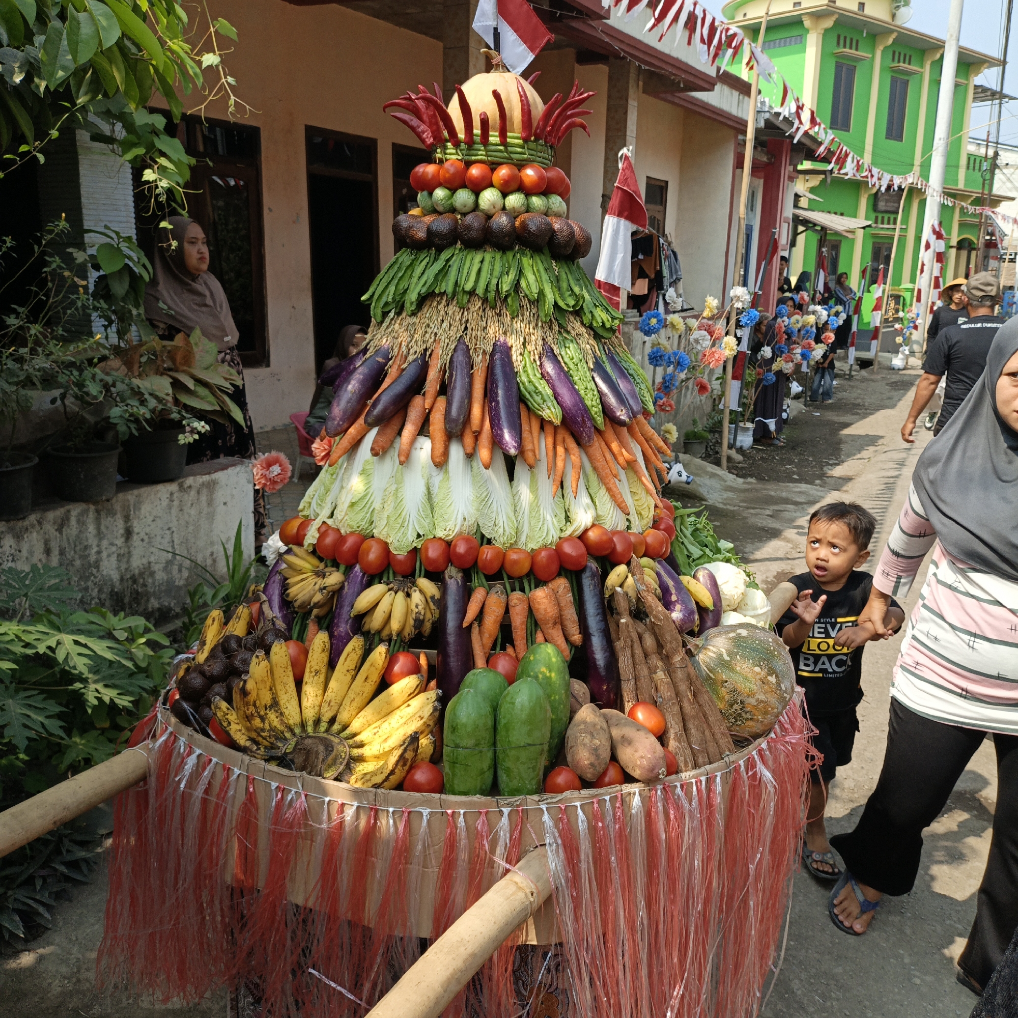 In Tanahbaya, Indonesia, a large, tiered display of fruits and vegetables stands prominently in an outdoor setting. The produce is arranged in a cone-like structure, with various items such as bananas, avocados, tomatoes, eggplants, carrots, bitter gourds, cabbage, and a squash visible. Red and white decorative streamers adorn the base of the display, suggesting a festive occasion.
Several people are present. A woman in a brown hijab is standing to the left of the display, partially obscured by foliage. In the foreground, a young boy in a black t-shirt is looking up with an expression of surprise or awe. To his right, a woman wearing a grey hijab and a striped shirt is partially visible, holding the boy's hand. Other individuals, some wearing black shirts, are seen in the background, walking along a path lined with colorful artificial flowers and Indonesian flags. The environment appears to be a residential street, with buildings visible in the background, one of which is painted green and yellow. The scene is illuminated by natural sunlight.