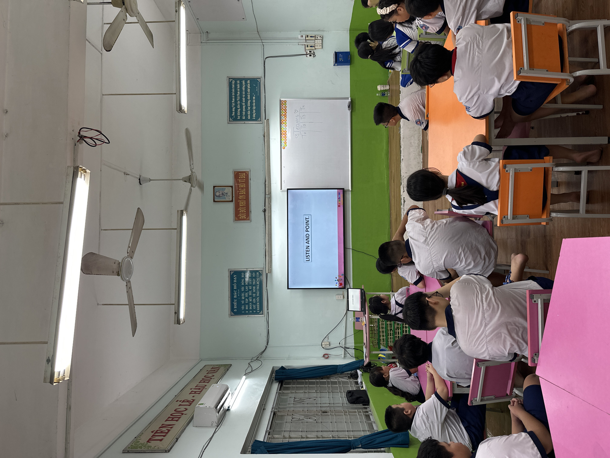 The image depicts a classroom in Ho Chi Minh City, Vietnam, where students wearing white shirts and dark blue shorts are sitting at tables facing a screen displaying the words "LISTEN AND POINT." The tables are arranged in rows with pink or orange tops. Some students are focused on the screen, while others appear to be interacting with each other. The classroom is illuminated by fluorescent lights and ceiling fans. A whiteboard is positioned behind the screen. Signs and posters with Vietnamese text are mounted on the walls. Windows with blue curtains are seen on the left side of the frame.