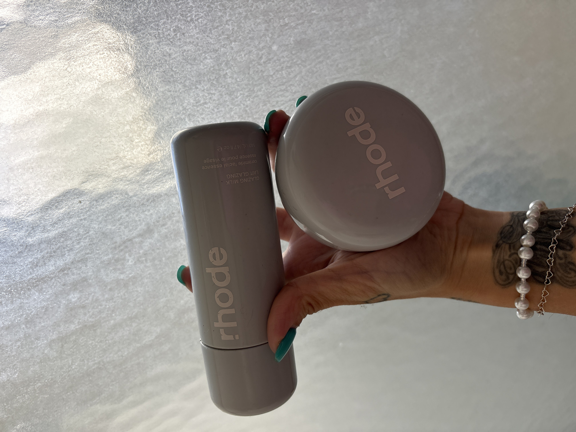 A hand with long, manicured green nails holds two items from the beauty brand "rhode". The hand wears a pearl bracelet and has visible tattoos. The background is a translucent, textured surface, possibly glass or plastic. The item on the left is a cylindrical bottle labeled "rhode GLAZING MILK LAIT GLAZING ceramide facial essence essence pour le visage 140JAL/4.7 fl oz e". The item on the right is a round container with the word "rhode" printed on top. The location, Seal Beach, United States of America, is not discernible from the image.