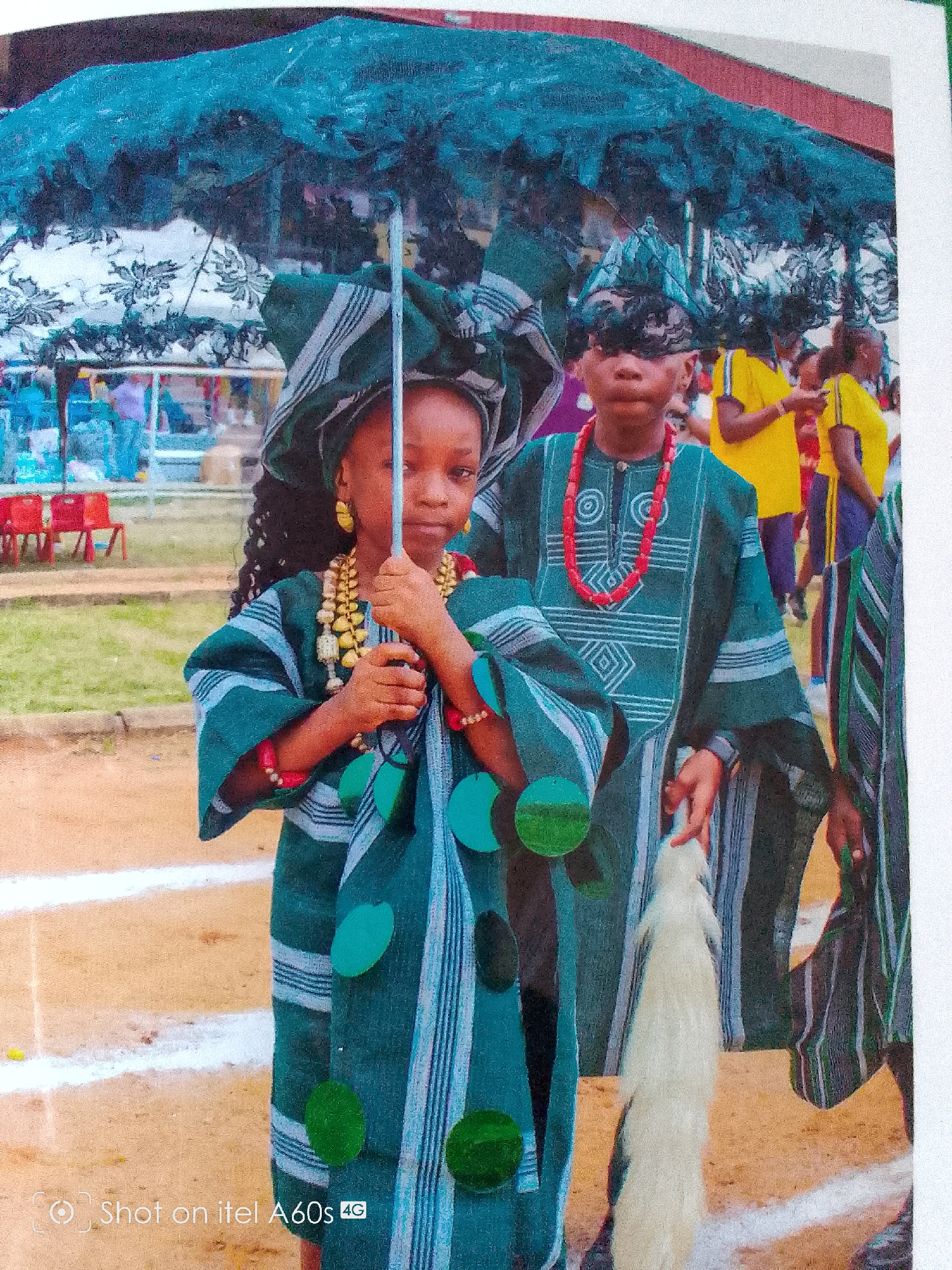 The image shows a group of children in Idimu, Nigeria, dressed in traditional attire. Two children are centrally located in the foreground. The girl in front is holding an umbrella with a silver shaft. She has dark hair, gold earrings, and beaded necklaces. The boy behind her is wearing a patterned green top and a red beaded necklace. Both are wearing matching headwraps. In the background, other people are visible, including individuals in yellow shirts. The setting appears to be outdoors with chairs in the background and what may be a running track on the ground. The bottom left corner of the image has the text "Shot on itel A60s 4G."