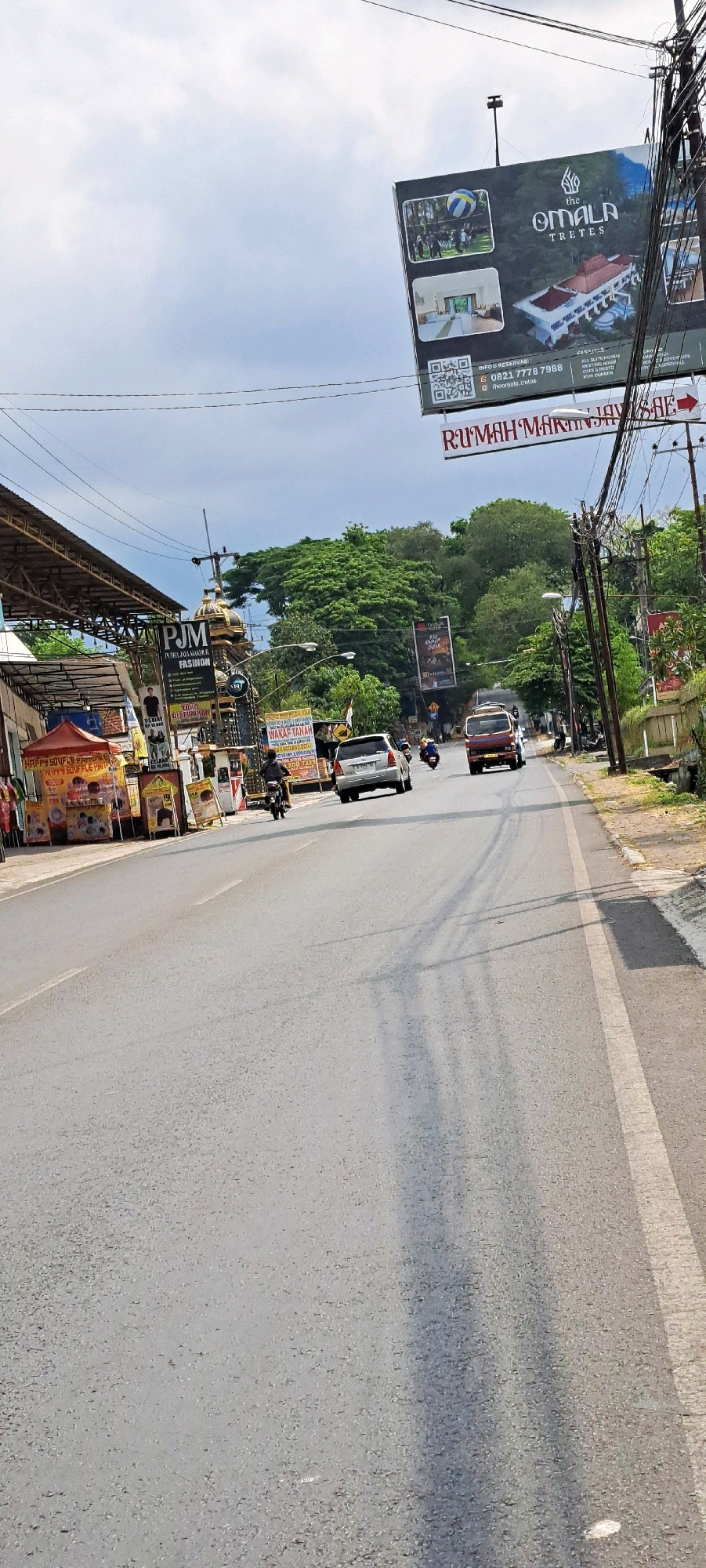 The image depicts a roadside scene in Pandaan, Indonesia, likely on a sunny day with scattered clouds. The foreground is dominated by an asphalt road with a white line marking the edge. On the left side of the road, there are several commercial stalls with awnings, displaying various products and signs, including "HAPPY SOUFFLE PANCAKE". Further down the road, a prominent building with a golden dome-like structure and "PJM FASHION" signage is visible.
To the right of the road, there are utility poles with numerous wires, and a large billboard advertises "THE OMALA TRETTES" with contact information and images of the resort. Another billboard further down reads "RUMAH MAKAN JAWA TIMUR".
Several vehicles are present on the road: a motorcycle with a rider in the foreground, a silver car, a red truck, and another motorcycle in the distance. People are visible on the motorcycles and near the stalls. The environment includes lush green trees in the background, suggesting a semi-urban or suburban setting. The overall impression is one of everyday activity along a busy street.