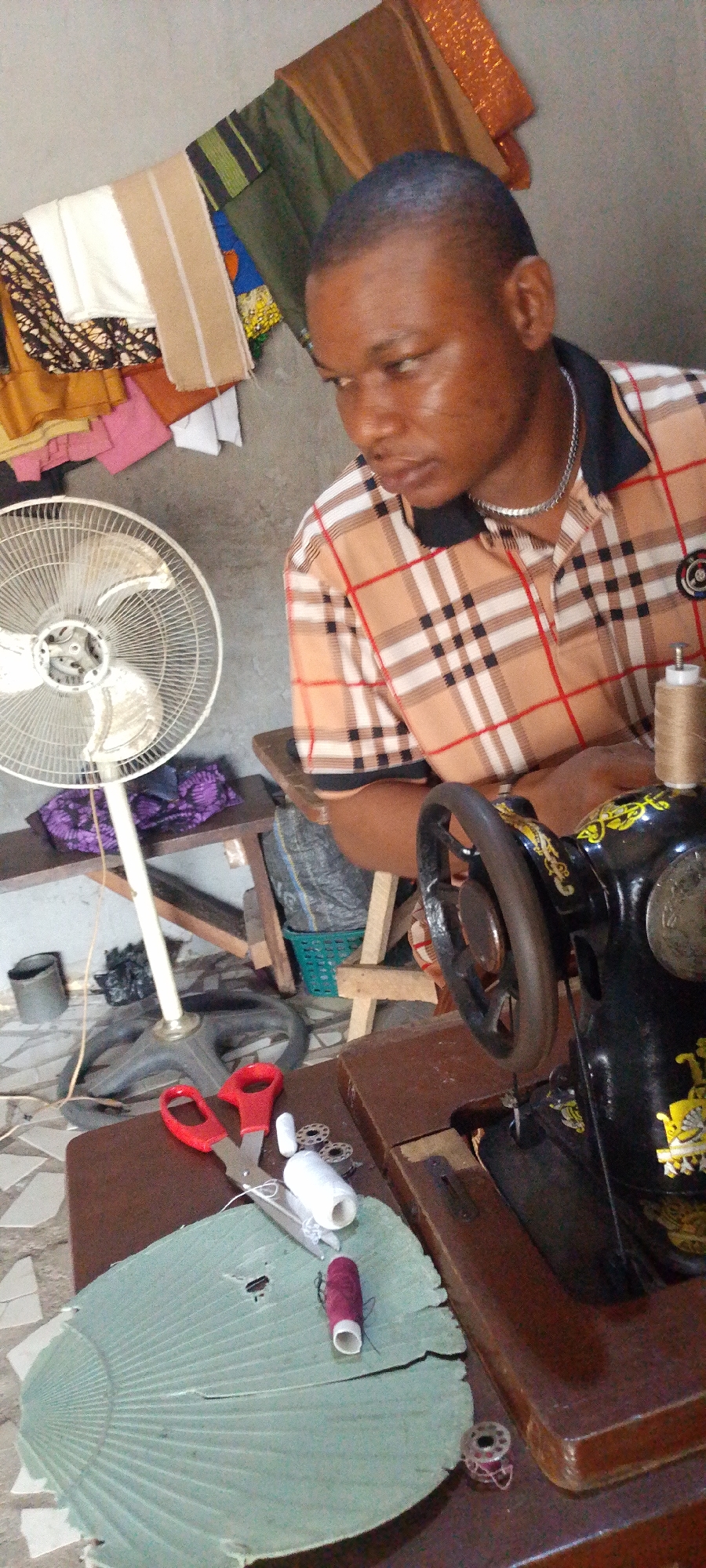 The image depicts a man seated at a sewing machine in Lokoja, Nigeria. The man, wearing a tan plaid shirt with a black collar and a silver necklace, is looking off to the side. He is positioned at an old black sewing machine with gold detailing. On the table next to the machine are red-handled scissors, spools of thread (white and burgundy), and several bobbins. A green fan lies on the table. A white electric fan stands to the left of the man. In the background, clothes are hanging on a wall. Other items visible include a wooden stool and a green wastebasket. The floor is tiled in a light color.