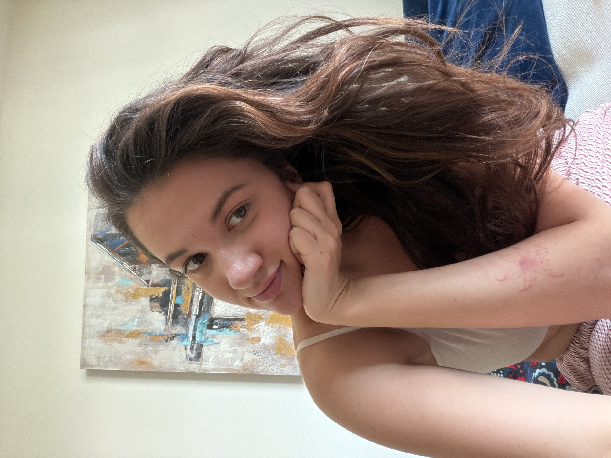 A young woman with long, wavy, brown hair is reclining indoors, likely on a sofa, as indicated by the blue, plush fabric behind her head and the patterned fabric partially visible at her waist. She is resting her head on her hands, positioned on her left side, and smiling slightly at the camera. She is wearing a white top with a thin strap. Her skin tone is fair. On her left arm, there is a faded, pink, floral-shaped mark, possibly a temporary tattoo or a scar. Behind her, on a light-colored wall, there is a framed abstract painting with shades of blue, brown, white, and gold. The location of the scene cannot be determined as being in Belgrade, Serbia, or any other location.