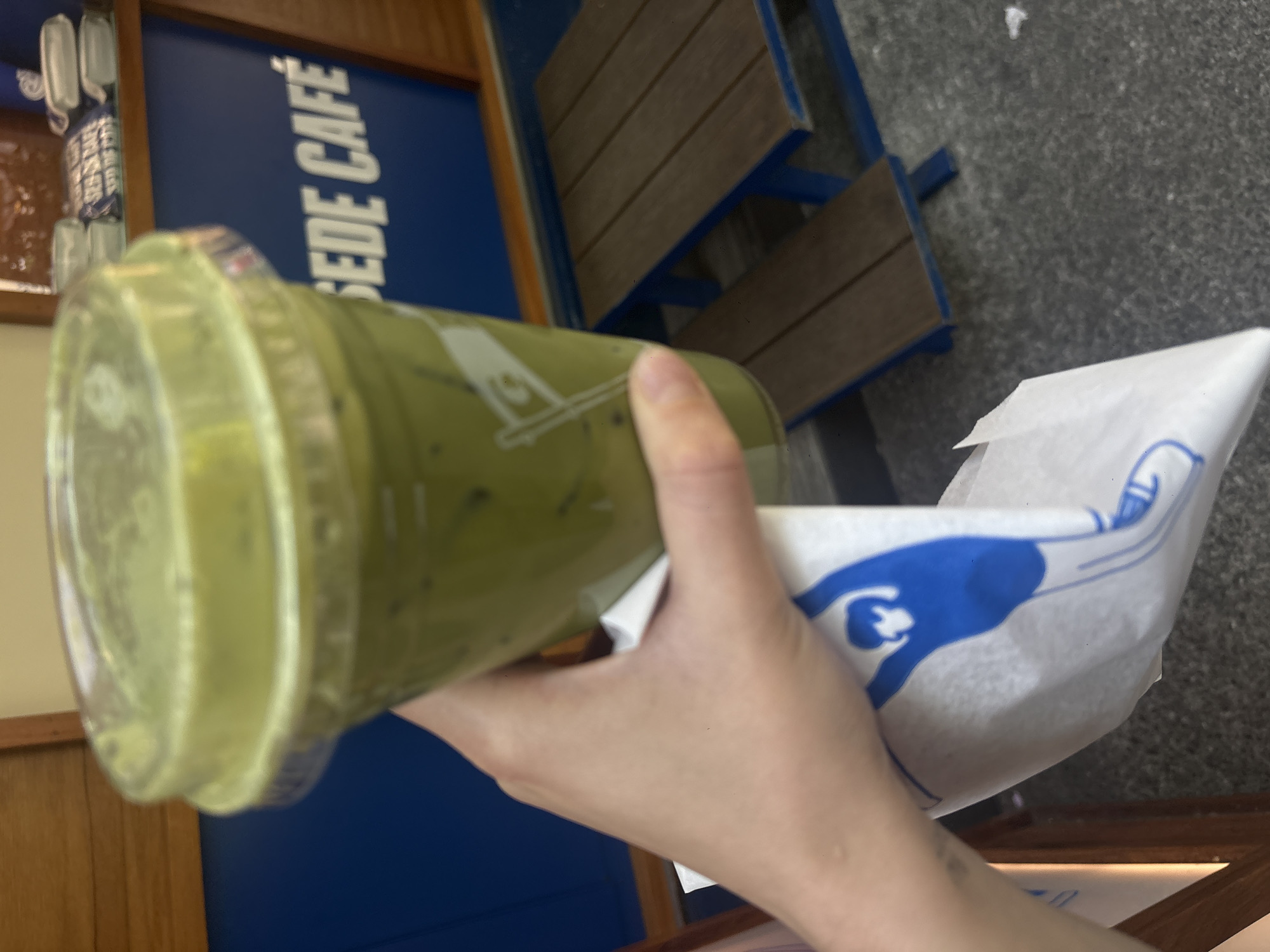 A hand holds a plastic cup filled with a green beverage and a white paper bag with a blue design. The cup's lid is translucent yellow-green, and the liquid inside appears to be a blend with dark speckles. The bag has an illustration in blue. The hand is pale with a slight tan, and a small tattoo is visible on the wrist. Behind the hand and cup is a blue wall with the words "SEDE CAFE" written in large, white, block letters. Above the wall is a wooden frame structure. In the background, there's a section of a picnic-style bench with wooden slats and blue supports, along with a gray concrete surface. The setting is in Mexico City, Mexico, suggested by the "Sede Cafe" branding.