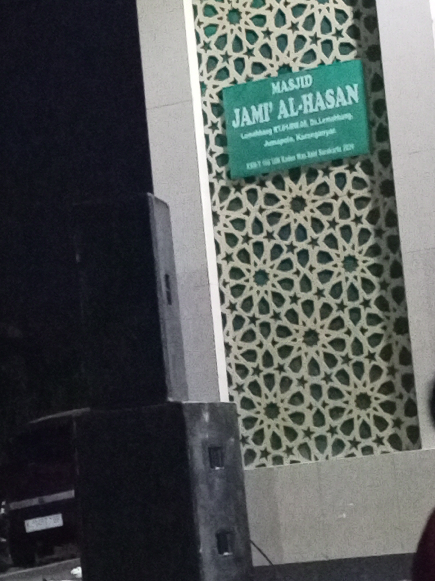 The image shows a street scene in Lemahbang, Indonesia. In the foreground, to the left, are two large black speakers. To the right, on a wall decorated with a geometric pattern, is a green sign with white text. The sign reads "MASJID JAMI' AL-HASAN" and includes smaller text below indicating "Lemahbang RT.1/RW.04, Ds.Lemahbang, Jumapolo, Karanganyar." There is also text at the bottom which appears to be "KMST 195 UN Raten Ras Asif Surakarta 2024". The wall behind the speakers is a light gray color. Further in the background, a dark-colored vehicle with a license plate is visible. The overall lighting suggests it may be daytime, but the scene is somewhat dark.