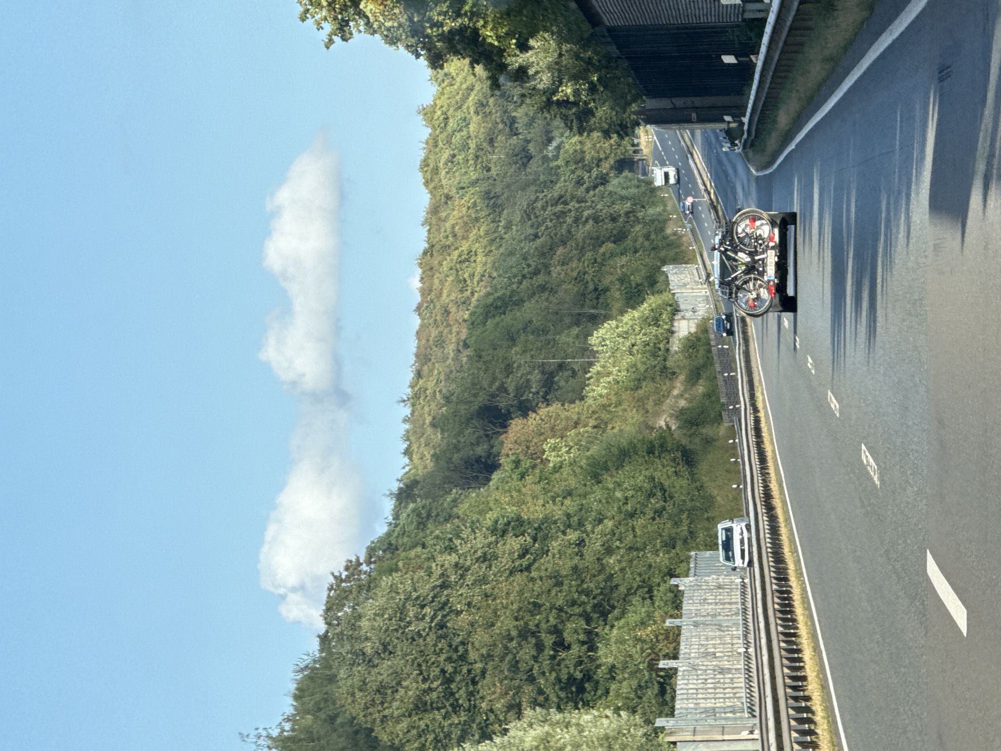 The image depicts a view from a vehicle traveling on a highway in Herzberg, Germany. The highway is bordered by dense green trees on both sides. The sky is blue with a few scattered clouds. In the foreground, a car is visible, carrying two bicycles on a rack at the rear. Further down the highway, other vehicles, including a campervan, are driving. The highway has multiple lanes and is marked with white lines. A sound barrier is present along the left side of the road. The scene appears to be a daytime, outdoor setting.