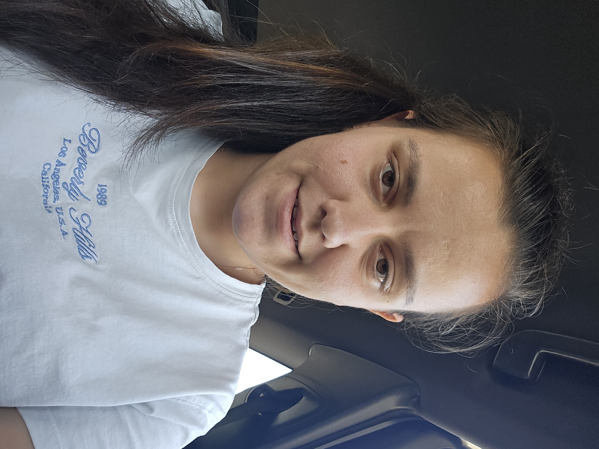 A woman with long, dark brown hair is pictured from the chest up, inside what appears to be a vehicle. She is wearing a light blue t-shirt with "1989 Beverly Hills Los Angeles, U.S.A. California" embroidered in blue script on the front. The woman is looking directly at the camera with a slight smile. Her face is slightly flushed, and her hair is pulled back into a ponytail on her left side. The background is dark, with a portion of the car's interior visible, including a sun visor and a window. The specific location in Staro Selo, Bulgaria, is not discernible from the image.