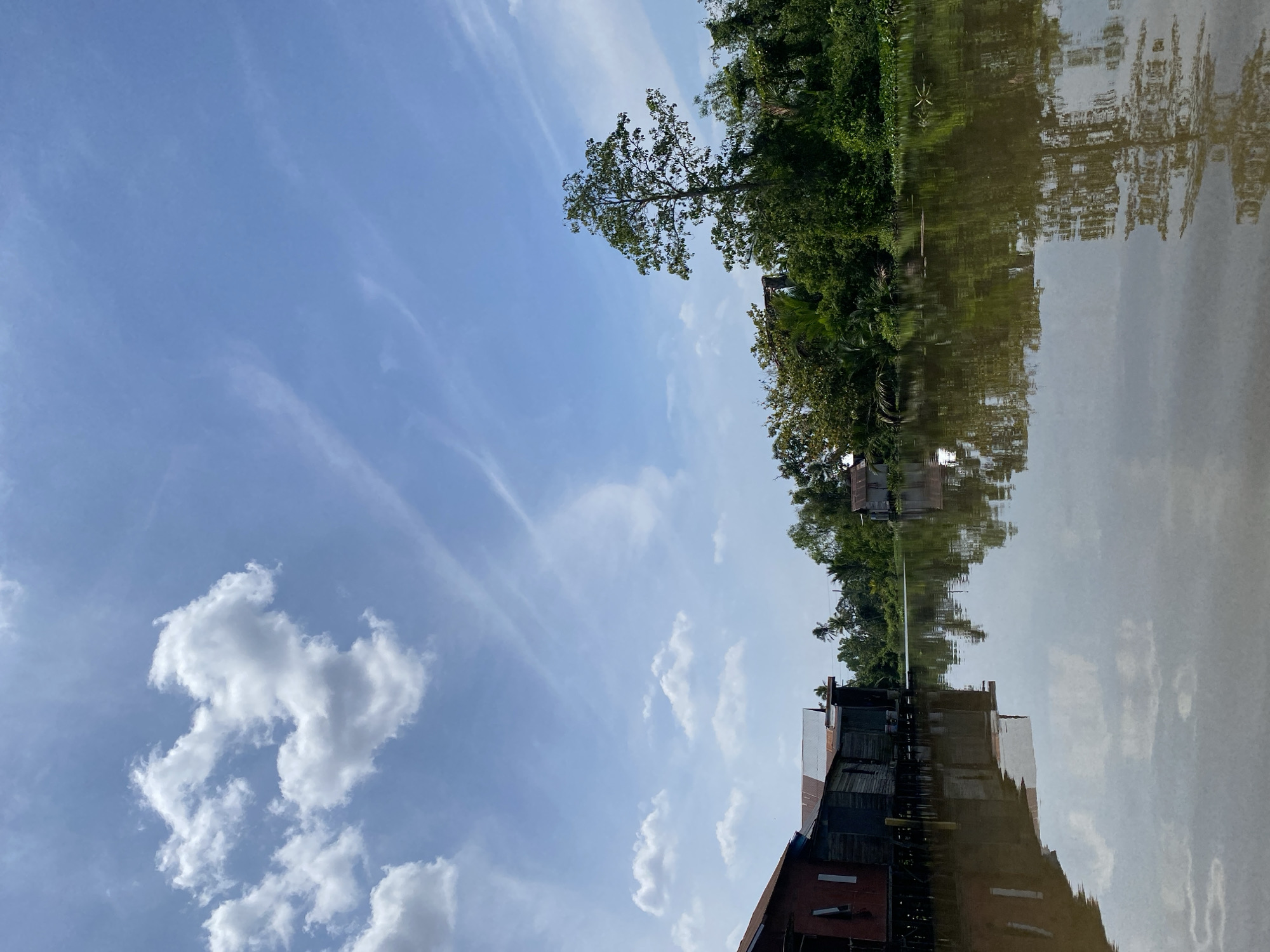 This is a horizontal, eye-level, wide-angle, outdoor, daytime photograph of the Martapura River in Banjarmasin, Indonesia. The river is wide and murky brown. On the left bank of the river are several stilt houses with weathered wooden walls and rusty metal roofs. The houses are clustered together and appear to be residential. The river reflects the houses and the sky. On the right bank, lush green vegetation, including trees and palm trees, grows thickly along the riverbank. A single, tall tree stands out against the background. The sky is a bright blue with scattered white clouds, some of which are wispy cirrus clouds. The overall impression is one of a tranquil, tropical riverside community.