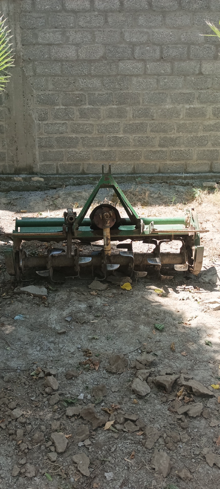 The image depicts a piece of agricultural equipment, specifically a rotary tiller, situated on a dirt and gravel surface. The tiller is green and shows signs of rust and wear. It is positioned in front of a cinder block wall. The ground is scattered with small rocks, debris, and some leaves. The scene appears to be outdoors, possibly on a farm or agricultural area. The location is Alam Khan Kalay, Pakistan.