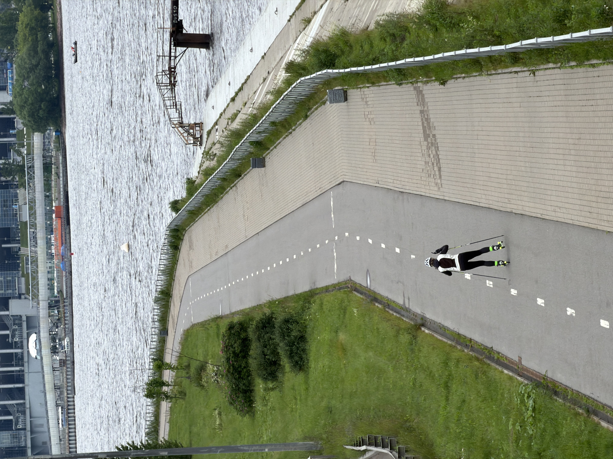 A person is rollerblading on a paved path alongside a body of water, with a view of city buildings and a bridge in the background. The person is wearing a white and black jacket, black pants, a white helmet, and bright green rollerblades. They are holding two ski poles. The paved path is grey with dashed white lines indicating lanes. To the left of the path is a grassy area with some bushes and trees. The body of water is choppy, suggesting wind or current. In the distance, a large modern building complex, possibly a stadium, is visible across the water, along with a bridge and some smaller boats. The scene appears to be in an urban park or recreational area in Saint Petersburg, Russia.
