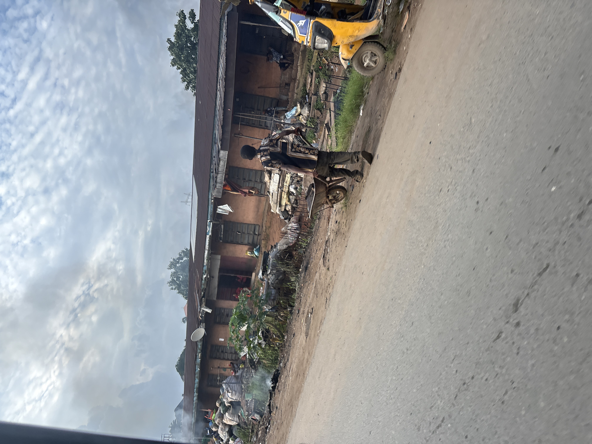 The image depicts a street scene in Onitsha, Nigeria, characterized by a dusty road, informal structures, and signs of economic activity. In the foreground, a person is pushing a wheelbarrow along the side of the road. The wheelbarrow appears to be carrying some items, though their nature is not clearly discernible.
To the right, a yellow auto-rickshaw, commonly known as a "Keke," is parked. These are ubiquitous modes of transportation in Nigeria. The background reveals a row of shops or market stalls with corrugated iron roofs. Piles of goods, possibly merchandise or refuse, are stacked near the structures. Smoke is visible in the air, suggesting an open fire or burning, which is a common practice for waste disposal or cooking in some urban areas of Nigeria. The overall impression is one of a bustling, albeit somewhat disorganized, urban environment. The sky is overcast with heavy clouds, indicating a possibly humid or rainy day.