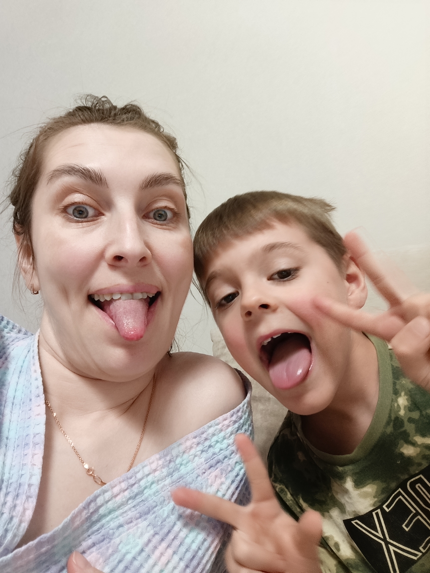 A woman and a young boy are taking a selfie indoors against a plain, light-colored wall. The woman is on the left side of the frame, wearing a light-colored, possibly tie-dye bathrobe that is slightly off her shoulder. She is sticking her tongue out and making a silly face, with wide eyes. She is also wearing a thin gold necklace. The young boy is on the right side of the frame, wearing a camouflage t-shirt with the word "REX" visible. He is also sticking his tongue out and making the peace sign with both hands. They are in close proximity, suggesting a close relationship. The location is not discernible from the image. There is nothing in the image to suggest the location is Chaikovsky, Russia.