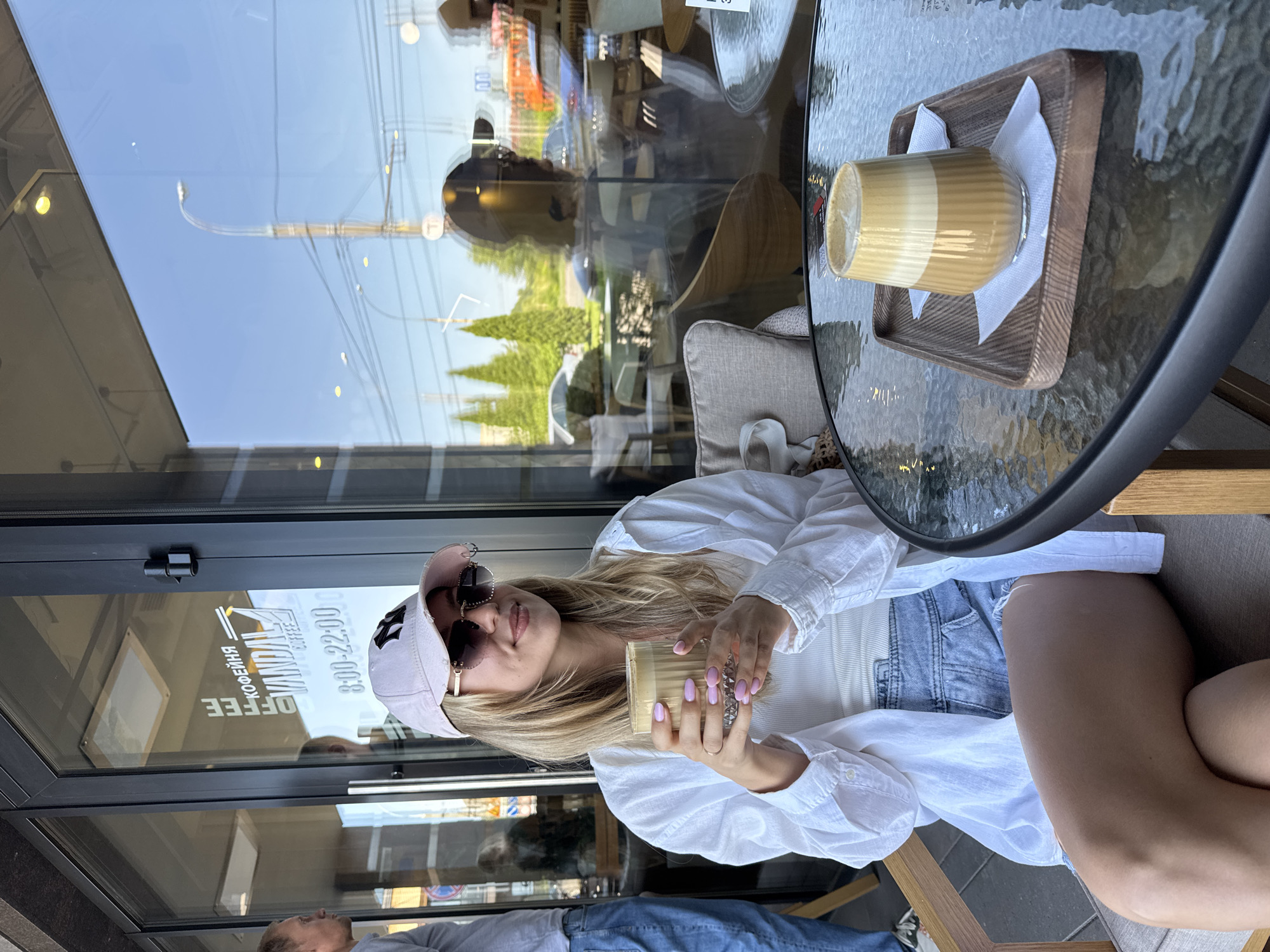 A young woman with blonde hair, wearing a white shirt, denim shorts, and a pink baseball cap, sits at a glass-topped table outside a cafe. She holds a glass of coffee in her hands, her nails painted a light purple color. The woman is looking at the camera with a slight smile. In the background, through the cafe window, reflections of trees and buildings are visible. A second coffee glass sits on a wooden tray on the table. The overall scene appears to be during the day, likely late morning or afternoon, with sunny weather. The text "VANDAL COFFEE" is visible on the cafe window, along with the hours "8:00-22:00".