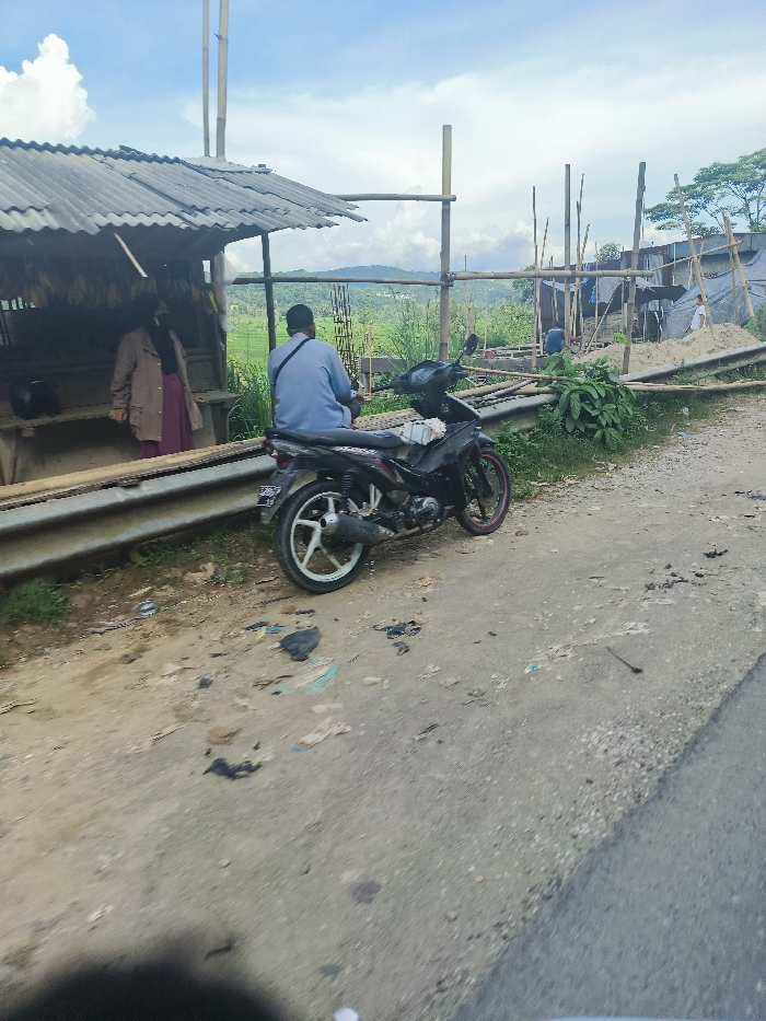 The image shows a man sitting on a black motorcycle on a dirt road in Sukatani, Indonesia, near the coordinates -6.631, 107.404. The man is wearing a blue shirt and is looking to the left. The motorcycle is parked on the side of the road, facing the right. There is a small building with a corrugated metal roof behind him, and another building further in the distance. There are green plants, including bushes and trees in the area. The road is lined with tall poles. There is a woman in a long red dress standing in the doorway of the building. There is debris and loose dirt scattered on the dirt road. There is a faint image of another vehicle in the foreground. The sky is blue and clear, with a few puffy clouds in the distance.