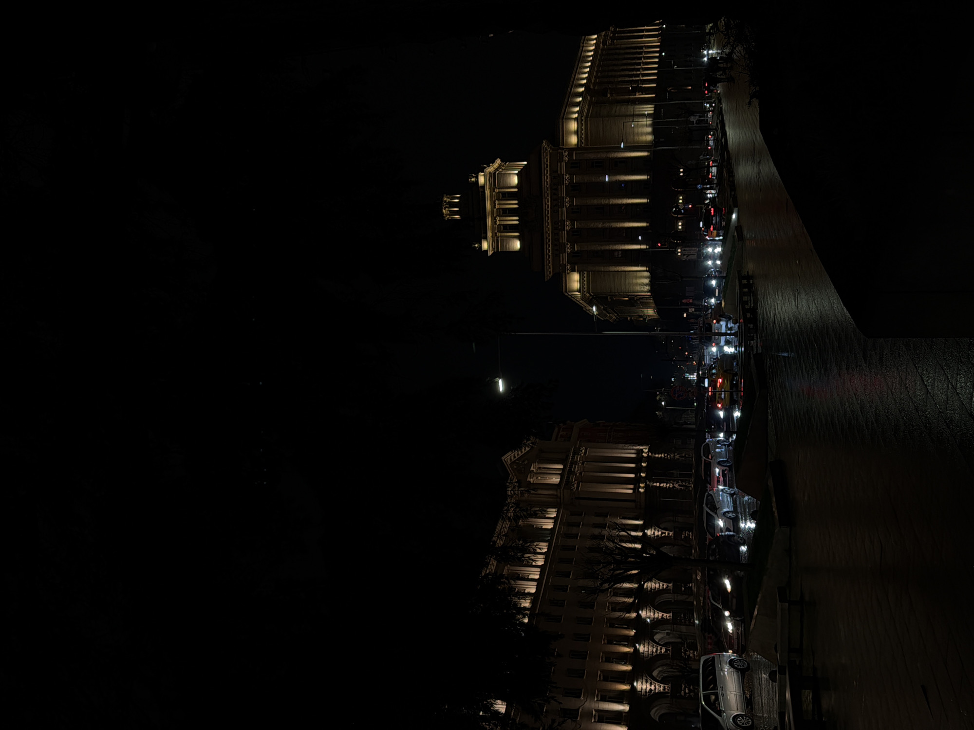 The image shows a night scene in Sofia, Bulgaria. The architectural style suggests the city center, possibly near Independence Square. Two large buildings with illuminated facades are visible, reflecting on a wet street. Cars line the street, their headlights adding to the city's ambiance. The sky is dark, with a faint glow from the moon or streetlights.