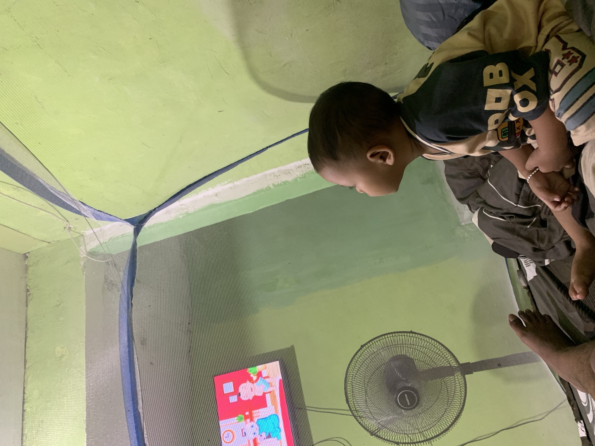 The image depicts a young child watching a television screen inside what appears to be a mosquito net enclosure in Palembang, Indonesia. The walls are painted green, and the floor is covered with bedding and a fan. The television displays a cartoon with two characters. The child is wearing a yellow and dark blue t-shirt and shorts, and a silver bracelet on their wrist. Part of an adult's foot is visible in the lower left corner of the image, suggesting the presence of at least one other person. The overall scene suggests a domestic setting, possibly a bedroom.