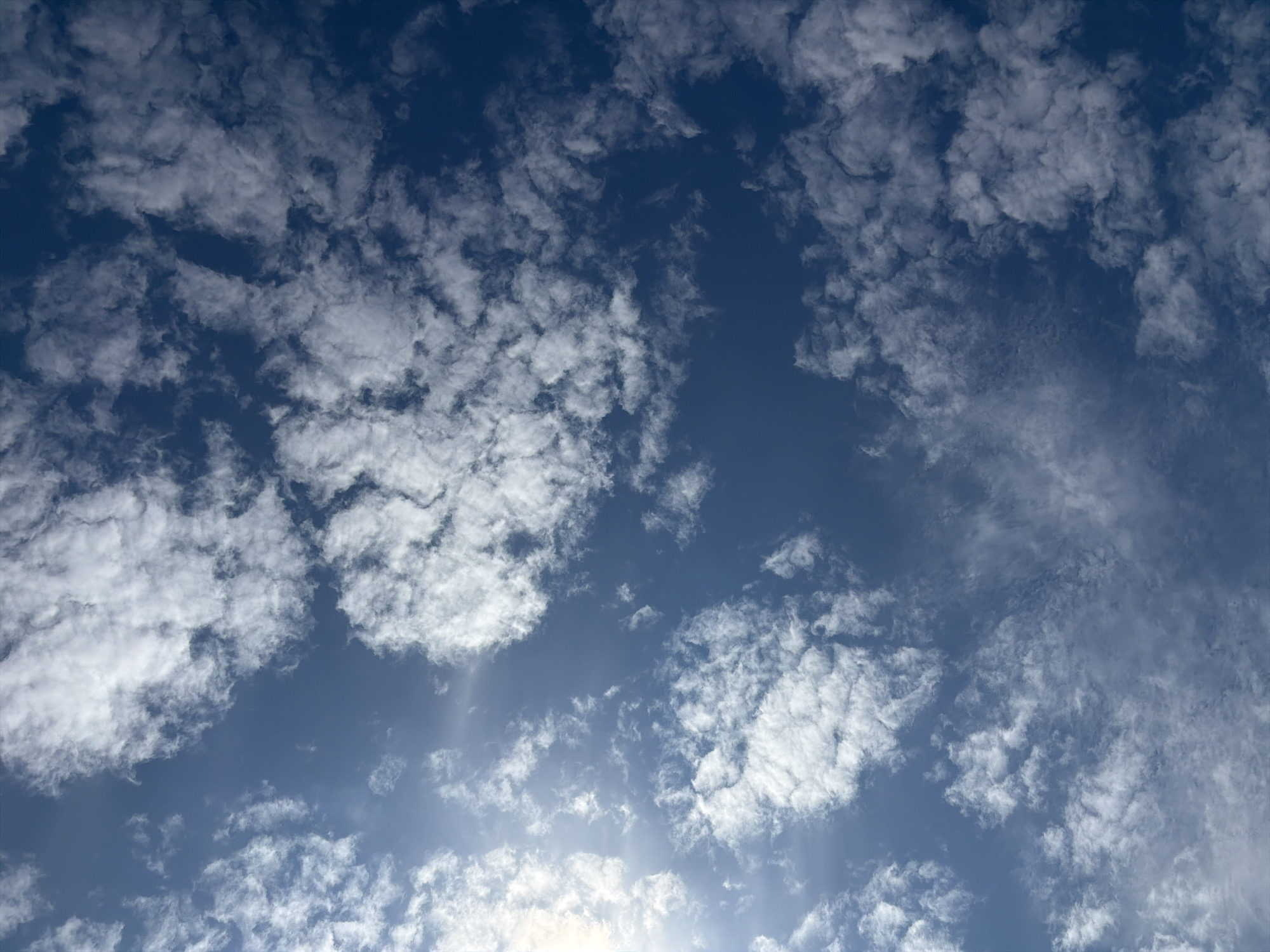 The image displays a vast expanse of a deep blue sky dotted with scattered white clouds of varying sizes and densities. The clouds appear wispy and fragmented in some areas, while others are more rounded and voluminous. Sunlight is visible, casting a bright glow from the upper left quadrant of the image, creating a slight lens flare effect. There are no visible structures, landforms, people, or any other discernible objects within the frame, making it a purely atmospheric scene. The location is specified as Oxentea, Moldova.