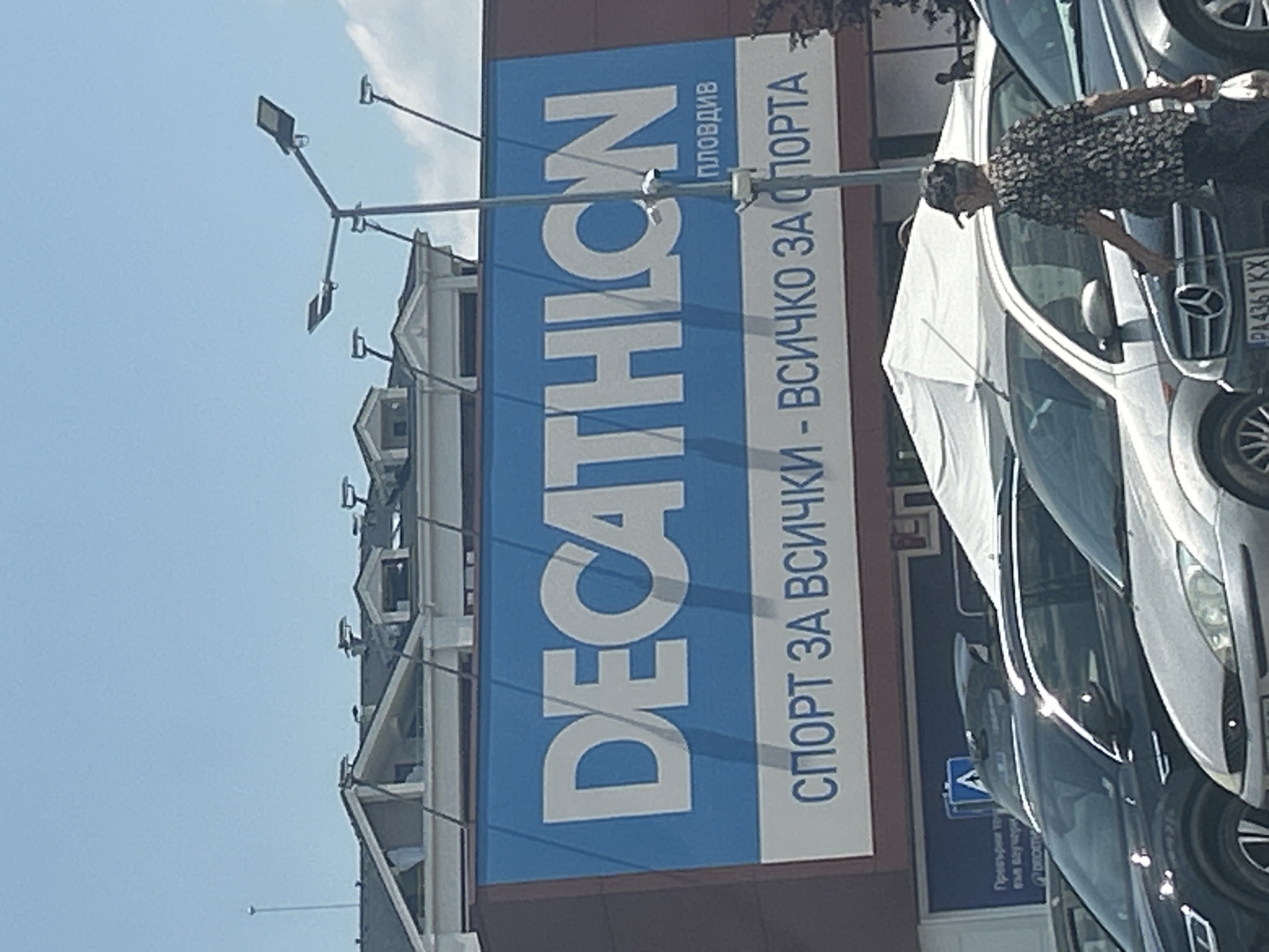 An outdoor medium shot captures a Decathlon store facade under a bright sky. The store's prominent blue sign displays "DECATHLON" in large white letters. Below this, in smaller white text, reads "СПОРТ ЗА ВСИЧКИ - ВСИЧКО ЗА СПОРТА" which translates to "SPORT FOR ALL - EVERYTHING FOR SPORT". To the right of "DECATHLON", the word "ПЛОВДИВ" (Plovdiv) is visible in smaller white text.
In the foreground, several cars are parked in a lot. A person, an elderly individual wearing a patterned shirt and a cap, is walking past a silver car, holding a plastic bag in their right hand and a cane in their left. The background shows the architecture of the Decathlon building with visible rooflines and dormer windows, and streetlights. Further back, another Decathlon store entrance is partially visible, with a blue sign featuring text in a foreign language. The overall scene suggests a sunny day in Plovdiv, Bulgaria.