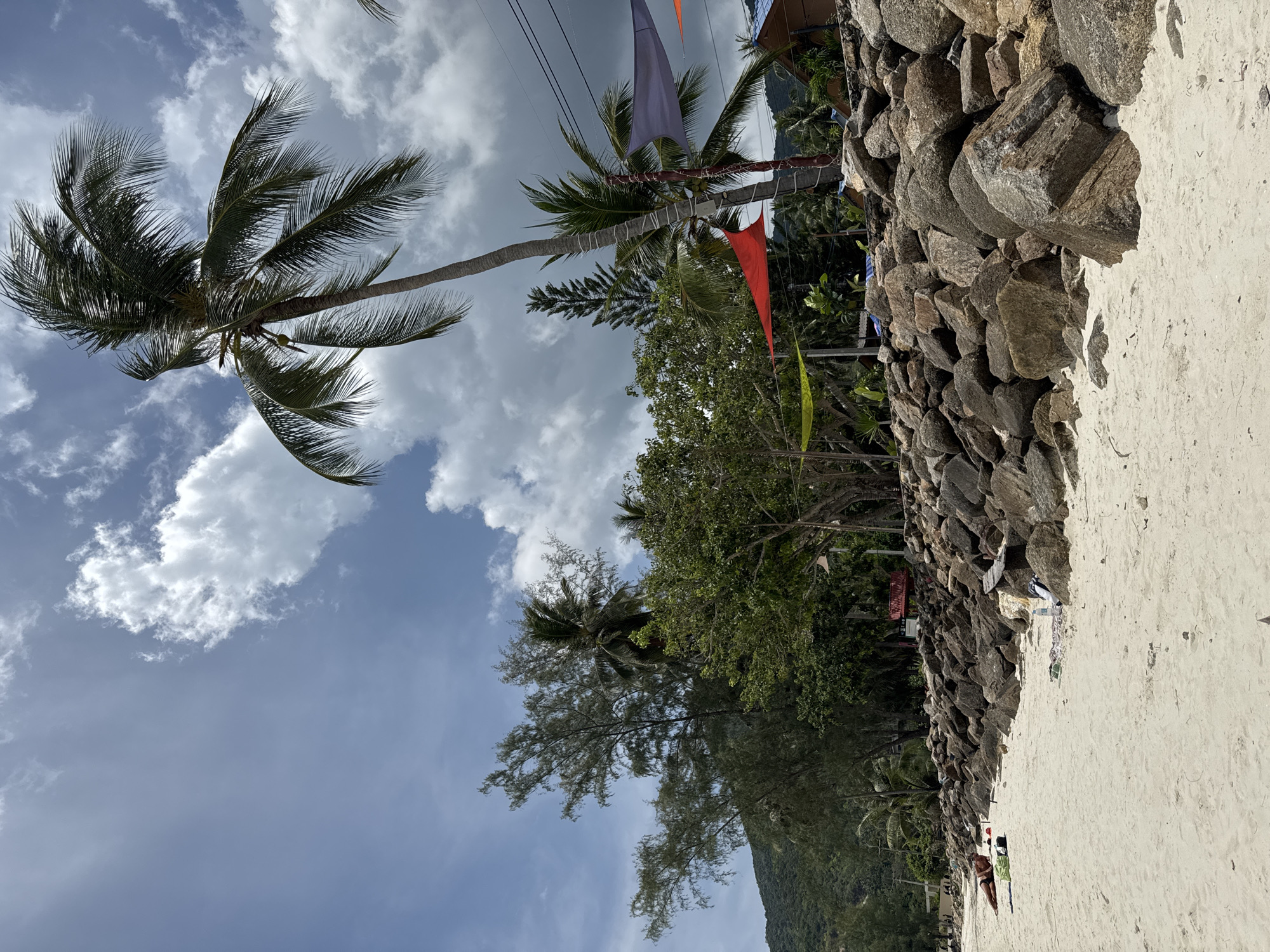 The image shows a beach scene in Baan Chaloklum, Thailand. A palm tree is on the left side of the image, with white sand in the foreground and a rock wall running along the middle right side of the scene. Lush trees and vegetation are positioned between the palm tree and rock wall and, further in the background, a glimpse of a mountain can be seen. Several colorful flags are strung between trees. The sky is bright with scattered clouds.