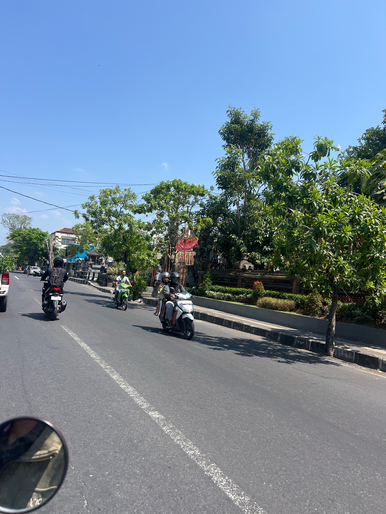 In Denpasar, Indonesia, on a sunny day, a street scene unfolds with several motorcycles on the asphalt road. A white motorcycle with two riders, a man and a woman, travels in the right lane. The man is driving, wearing a helmet, and the woman is seated behind him, also with a helmet. To their left, a green motorcycle with a single rider, wearing a helmet, moves in the same direction. Further ahead, a black motorcycle with a rider wearing a backpack and helmet is in the left lane. The road is marked with a white dashed line. On the right side of the road, there is a sidewalk with a stone wall and lush green trees and foliage. Buildings are visible in the background. The sky is clear blue. The image appears to be taken from the perspective of someone on a motorcycle.