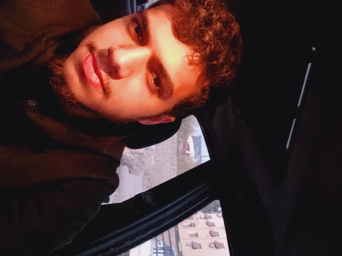 The image depicts a young adult male situated within the dark interior of a vehicle, appearing to take a self-portrait. He has dark, curly hair, a beard, and wears a dark, collared jacket with a zipper. His gaze is directed forward, and his tongue is slightly visible between his lips. A strong light source from the front-right illuminates the right side of his face and upper body, while the left side remains in shadow. Through the vehicle's windows, an urban exterior of Kafr Sousa Municipality, Syria, is visible. On the right, a multi-story, light-colored building with multiple windows and several protruding exterior units, possibly air conditioning units, is present. Further in the background, additional structures and a light-colored wall or rocky formation can be discerned. The vehicle's dark overhead console is partially visible at the top of the frame.