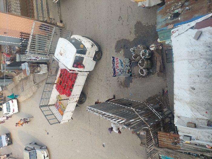A truck filled with red baskets sits on a street, alongside a small white van. Adjacent to them are multiple woven baskets, possibly containing goods for sale. A sign stands nearby. The street scene includes several cars and pedestrians. Overhanging structures and buildings border the road, creating an urban setting. The image appears to be taken from above, possibly from a higher building.
