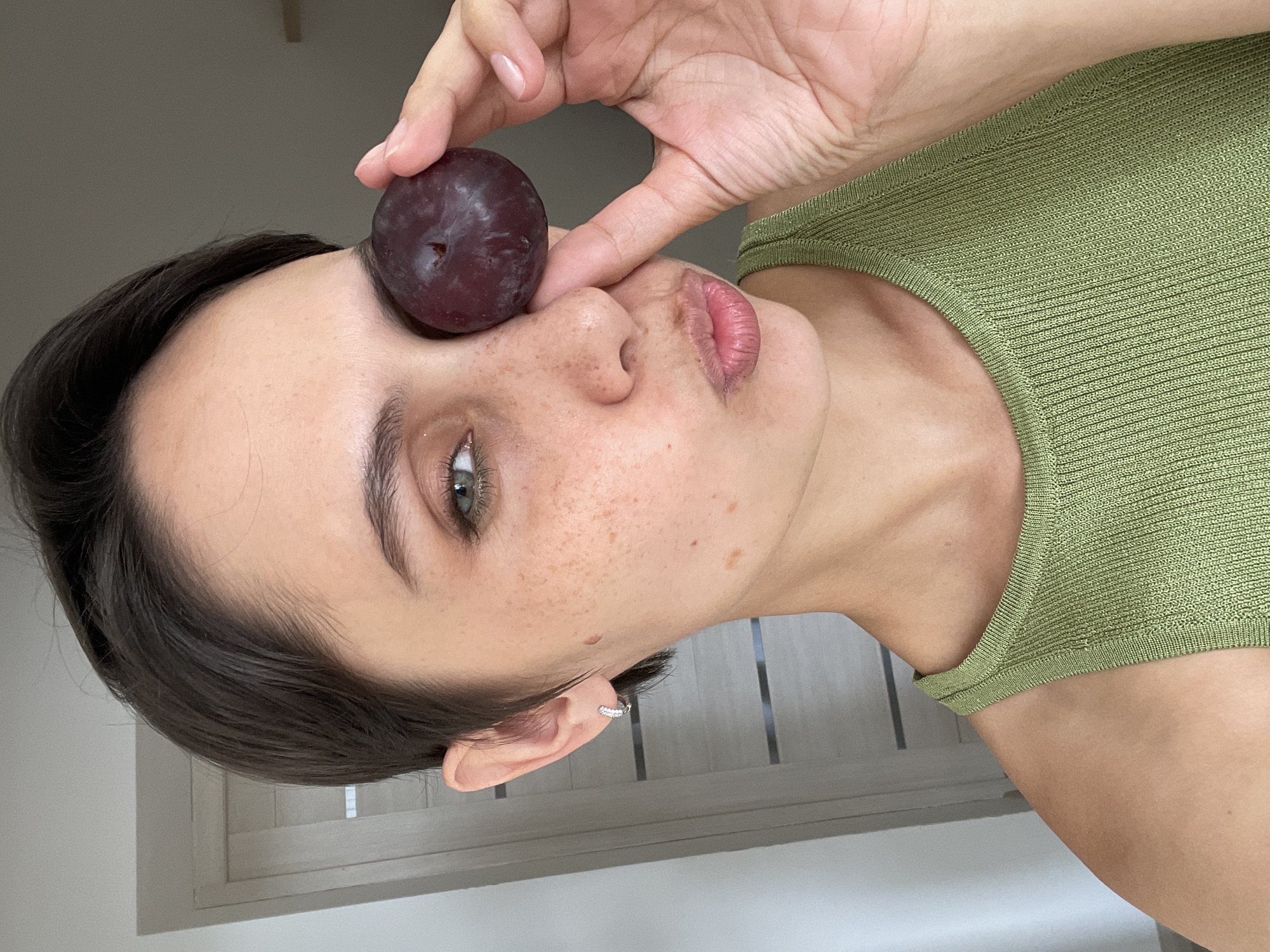 A woman is holding a plum against her eye. She has short, dark hair and fair skin with freckles. She's wearing a light green top. Her ear has a small, silver hoop earring. The background is a room with a white door and wall. The location is not specified.