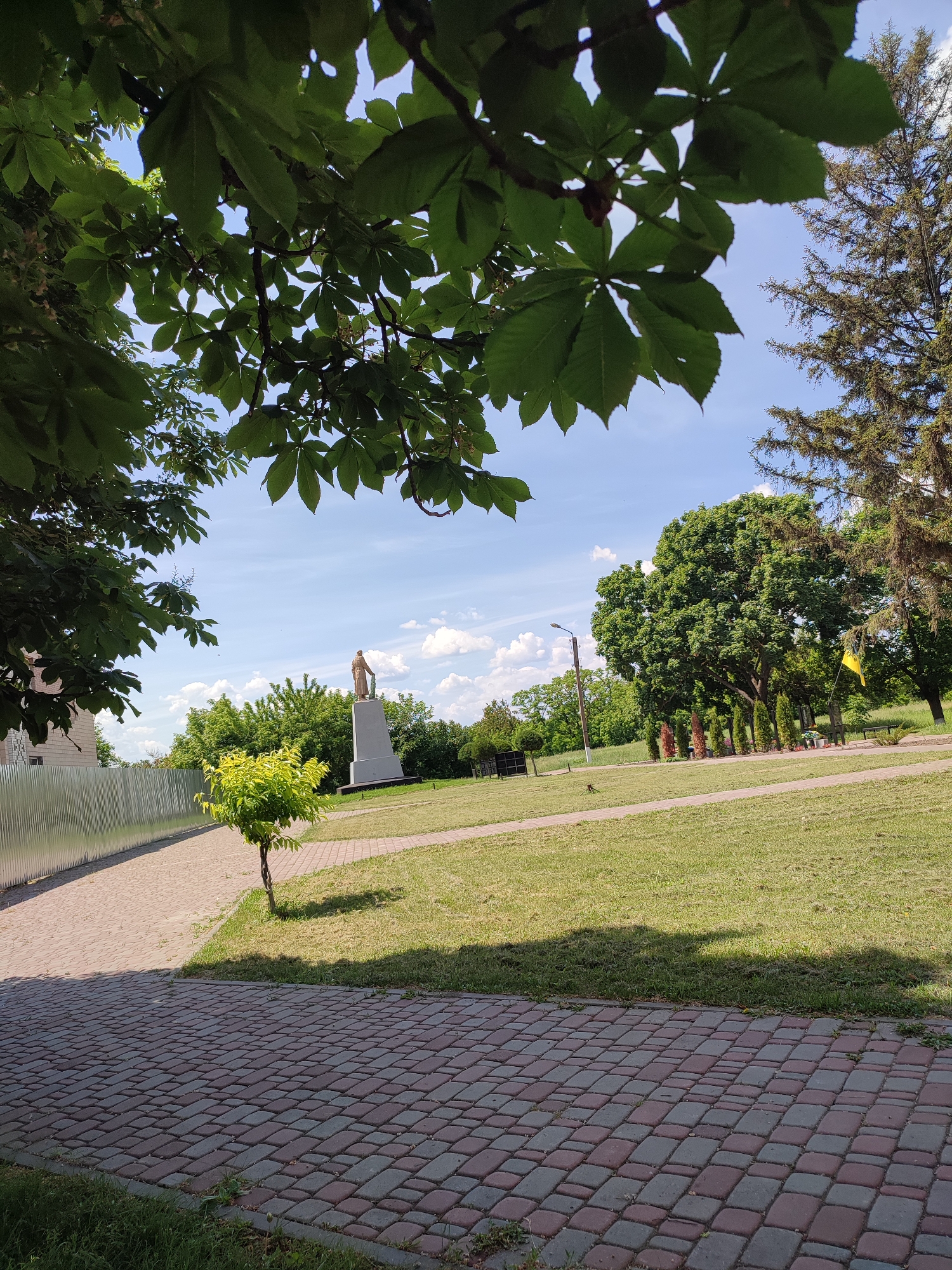 The image captures an outdoor scene, likely a public park or green space, on a clear, sunny day in the city of Unknown, Unknown. The perspective is low-angle, looking up and out from beneath the canopy of a large tree, whose dark green, palmate leaves (consistent with a horse chestnut) dominate the top third of the frame. Sunlight filters through these leaves, casting shadows and highlights.
In the foreground, a paved pathway made of interlocking reddish-brown and grey pavers extends across the bottom of the image. Shadows, presumably from the overhead foliage, fall across this path and adjacent mown grass. To the left, another lighter-colored paved path runs parallel to a tall, corrugated metal fence. A small, bright green-leafed tree with a slender trunk stands in the grassy area between these two paths.
The midground features a large expanse of mown grass, intersected by a curving paved path similar to the one in the foreground. A prominent light-colored statue of a human figure, possibly male and appearing to hold an object like a sword, stands atop a tall, grey, rectangular pedestal. Behind the statue, a dense line of diverse green trees forms a natural boundary across the scene. On the far right, partially obscured by trees, a yellow flag is visible on a pole. A tall, dark-trunked coniferous tree stands prominently on the upper right.
The background reveals a bright blue sky with scattered white cumulus clouds. Additional distant trees and greenery are visible, suggesting an expansive natural area. No people are visible in the scene.