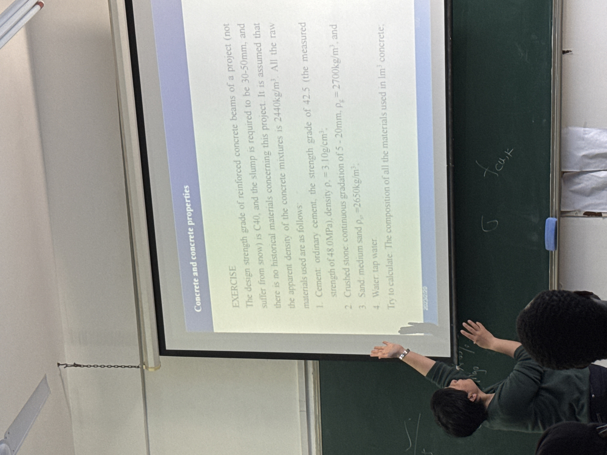 An indoor, slightly tilted medium shot captures a classroom or lecture setting, likely in Zhongtun, China. The focal point is a projector screen displaying an exercise related to "Concrete and concrete properties." The text on the screen details a problem about reinforced concrete beams, specifying design strength (C40), slump (30-50mm), apparent density of concrete mixtures (2440kg/m³), and properties of raw materials: ordinary cement (strength 42.5/48.0MPa, density 3.10g/cm³), crushed stone (5-20mm, density 2700kg/m³), medium sand (density 2650kg/m³), and tap water. The task is to calculate the composition of materials for 1m³ of concrete. A date, "2023/2/20," is visible at the bottom of the slide.
To the right of the screen is a dark green blackboard. A person with dark hair, wearing a dark green long-sleeved top, is standing in front of the blackboard, with their arms outstretched, seemingly explaining or gesturing towards it. Some handwritten text is visible on the blackboard, including "G" and "fcu,k." Part of another person's head with curly dark hair is visible in the lower right foreground. A white ceiling fan is partially visible in the upper left corner of the room. The overall lighting suggests an indoor, well-lit environment.