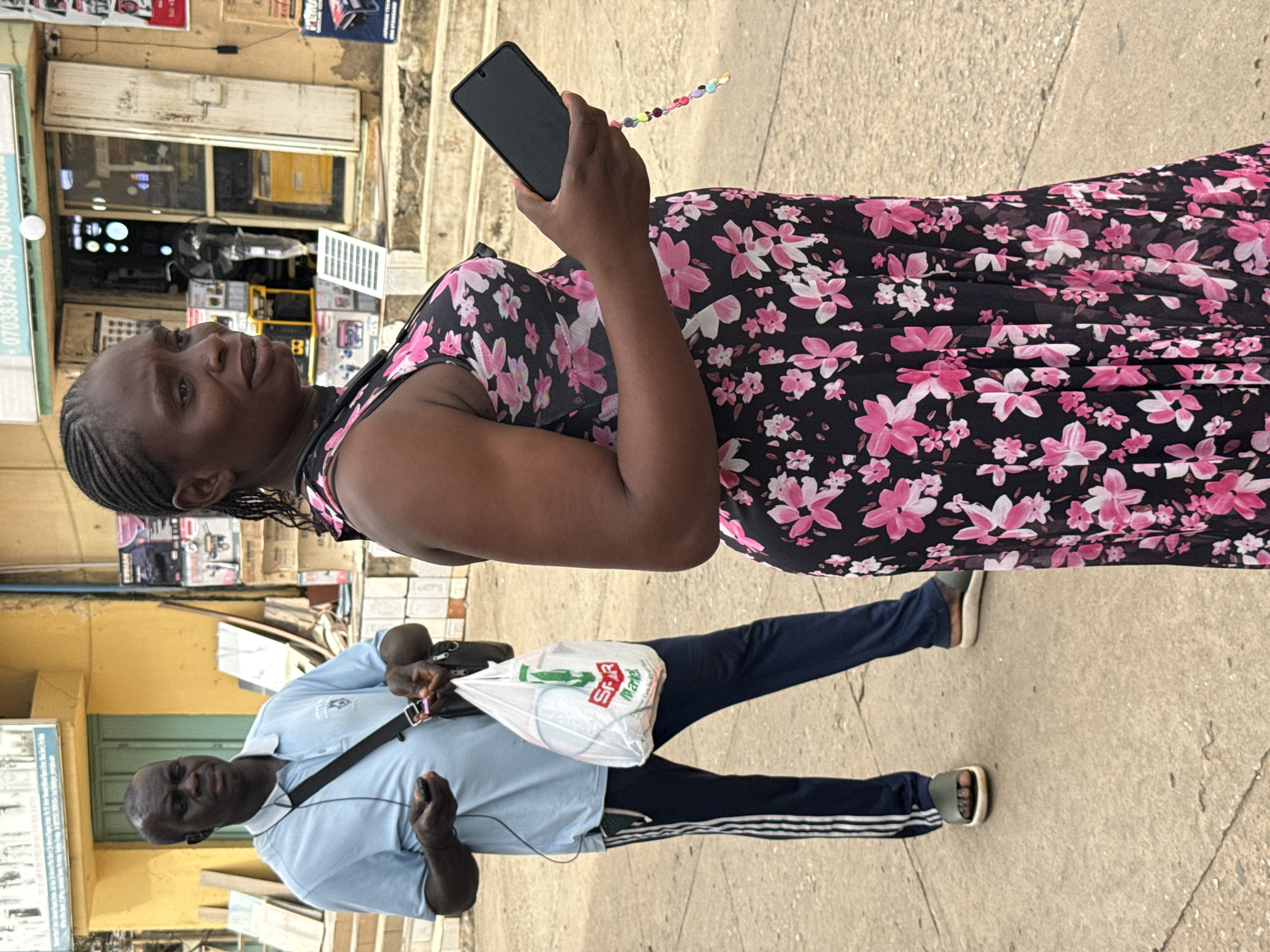An outdoor, medium shot captures two individuals in Abuja, Nigeria, standing on a paved walkway adjacent to a storefront. In the foreground, a woman with braided hair wears a sleeveless black dress adorned with a pink floral print. She holds a black smartphone in her right hand, from which dangles a colorful beaded strap. To her left, a man in a light blue polo shirt and dark track pants with white stripes carries a white plastic shopping bag from "SPAR Market" in his left hand and a dark object, possibly a phone, in his right. He also has earphones connected to his ears and a dark bag slung across his body. The background reveals a yellow building with a green doorway and a storefront displaying various appliances and goods, including what appear to be generators. The scene suggests a commercial area in the city.