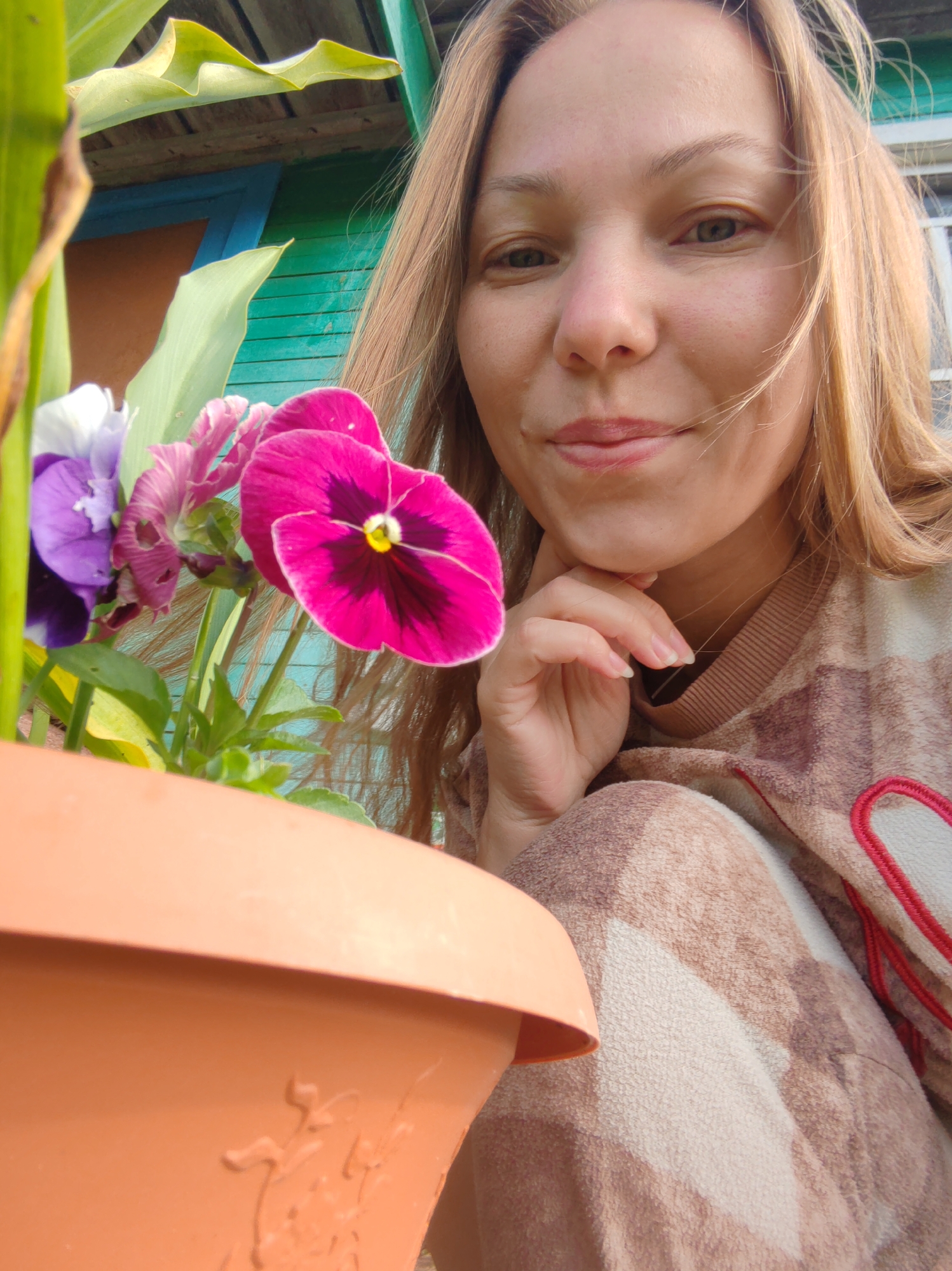 A woman with blonde hair is captured in a close-up selfie, her face turned towards the camera with a slight smile. She rests her chin on her hand. In the foreground, a terracotta pot contains vibrant pink and purple pansies, with large green leaves visible to the left. The background shows a weathered teal wooden structure with orange and blue accents, suggesting an outdoor setting likely in a rural or suburban area, possibly in the city of Чухлэм, Russia, given the context. She is wearing a patterned, fuzzy sweater in shades of brown and white. The overall lighting is natural and bright.