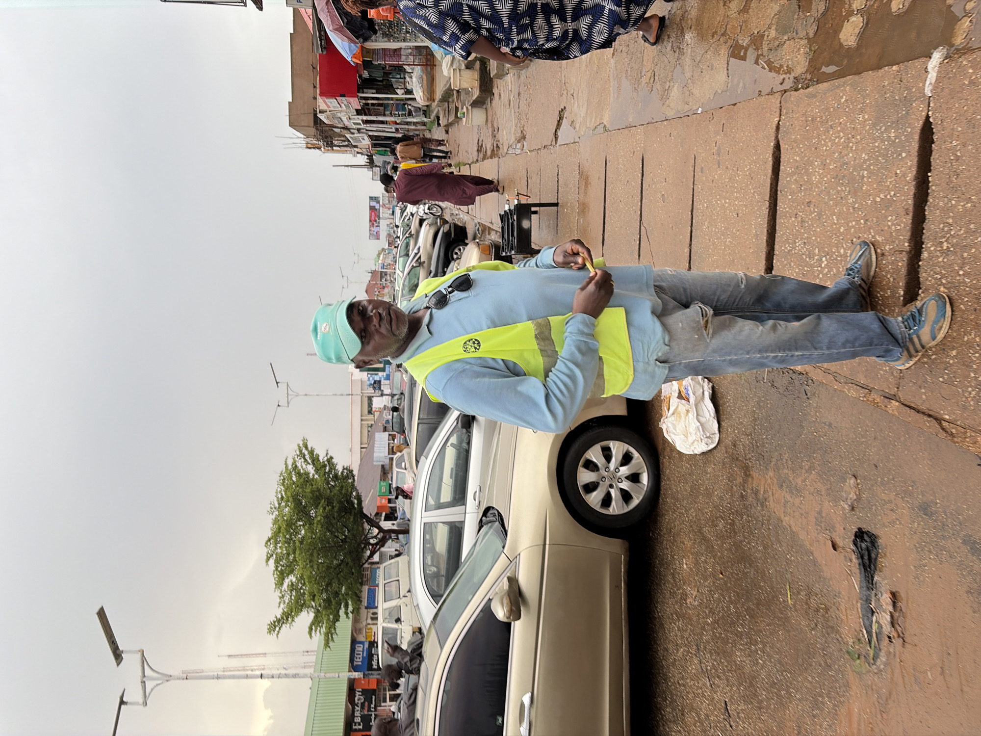 A man stands in the foreground on a sidewalk in what appears to be a commercial area of Jos, Nigeria. He is wearing a light blue long-sleeved shirt, a bright yellow safety vest, blue jeans with rips, and athletic shoes. He has a light green baseball cap on and sunglasses pushed up on his head. He is holding some small yellow items in his right hand. Behind him, a street is lined with cars parked along the side. The ground is wet and appears to be muddy, with puddles of water present, suggesting recent rain. In the background, there are buildings with shops and signs, as well as streetlights and trees. Other people are visible walking along the sidewalk and in the street. The sky is overcast.