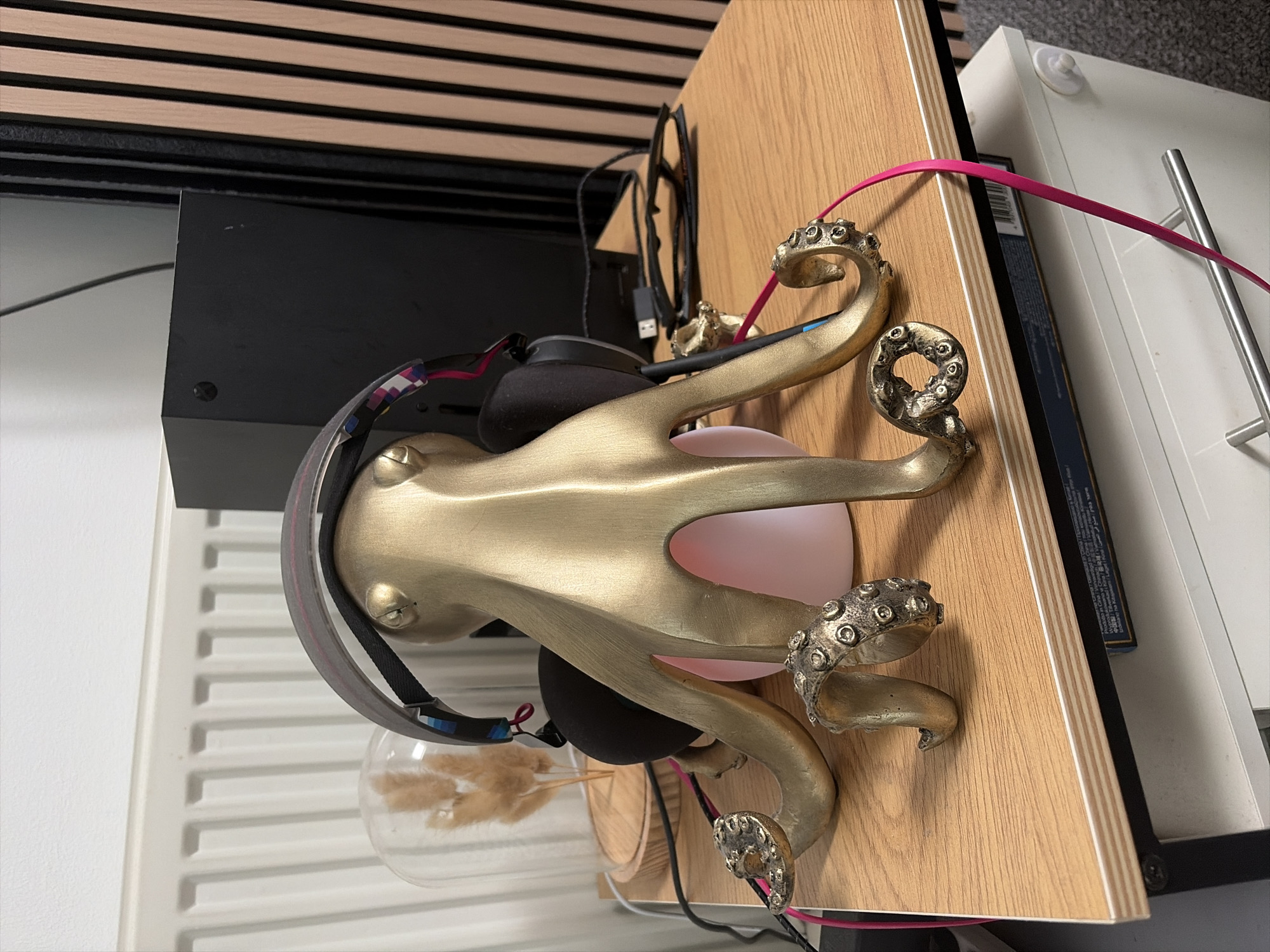 The image shows a golden octopus sculpture perched on a light wood-grained surface. The sculpture is holding up a pair of black headphones with a pink and gray headband. Underneath the headphones is a round glass vase containing dried plant material. Behind the octopus, to the left, is a black rectangular object and a white radiator. Various black cables are visible, along with a pink cable extending to the right of the sculpture. The location is Salford, United Kingdom.