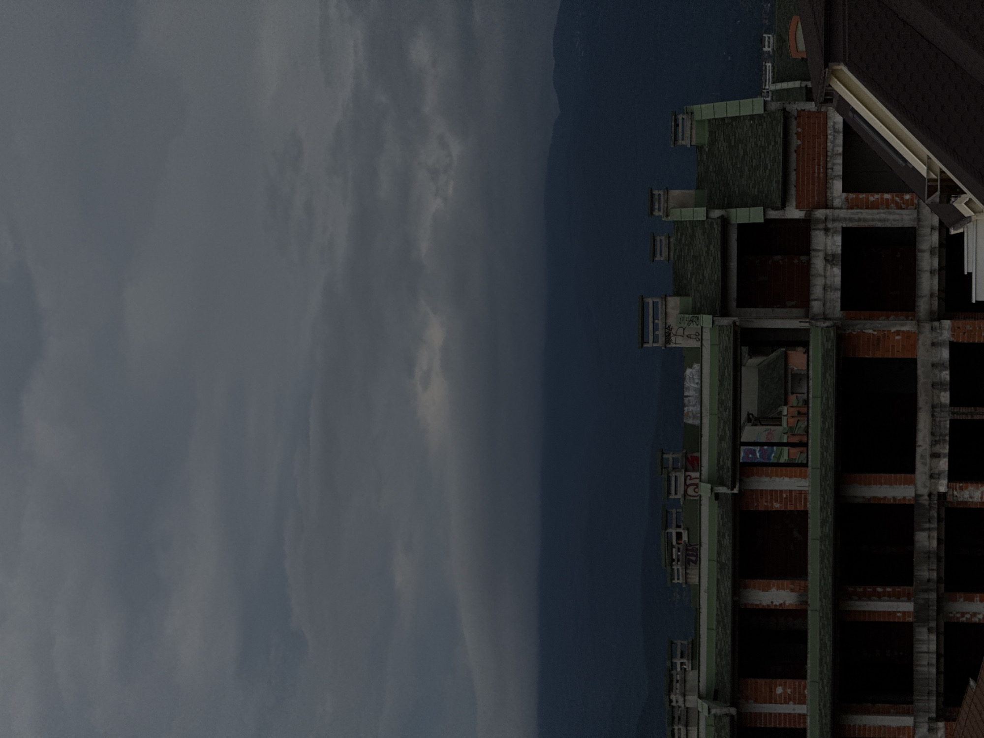 The image depicts the construction of a multi-story building under a dark, cloudy sky. The building appears to be unfinished, with exposed brickwork and dark window openings. Several chimneys or ventilation shafts are visible on the roof, some with graffiti. In the background, dark mountains rise against the overcast sky. The scene suggests a possibly abandoned or stalled construction project. There are no people visible in the image.