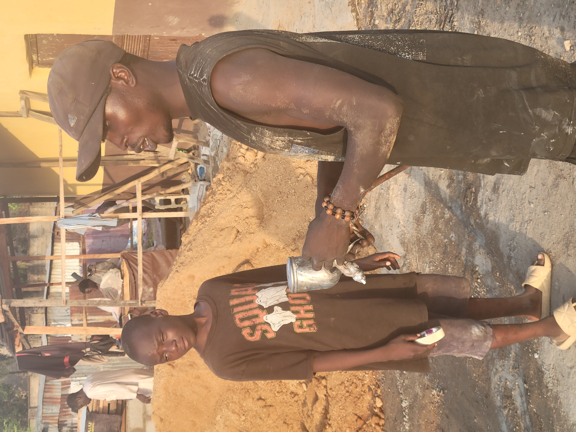 The scene, likely in Kwamba, Nigeria, shows two individuals near a pile of earth or construction material. One individual, possibly a child, wears a brown t-shirt with a "Ghost" graphic, gray shorts, and light-colored sandals. They hold a small white object in their left hand, and an older person, wearing a black sleeveless shirt, a dark baseball cap, and beaded bracelets, is holding a metallic object over them. Dust or particulate matter covers their arms and upper torso. In the background, there appears to be a partially constructed or dilapidated building with corrugated metal sheets. Two other people stand in the background. The overall environment appears to be outdoors with a mix of construction debris and signs of activity.