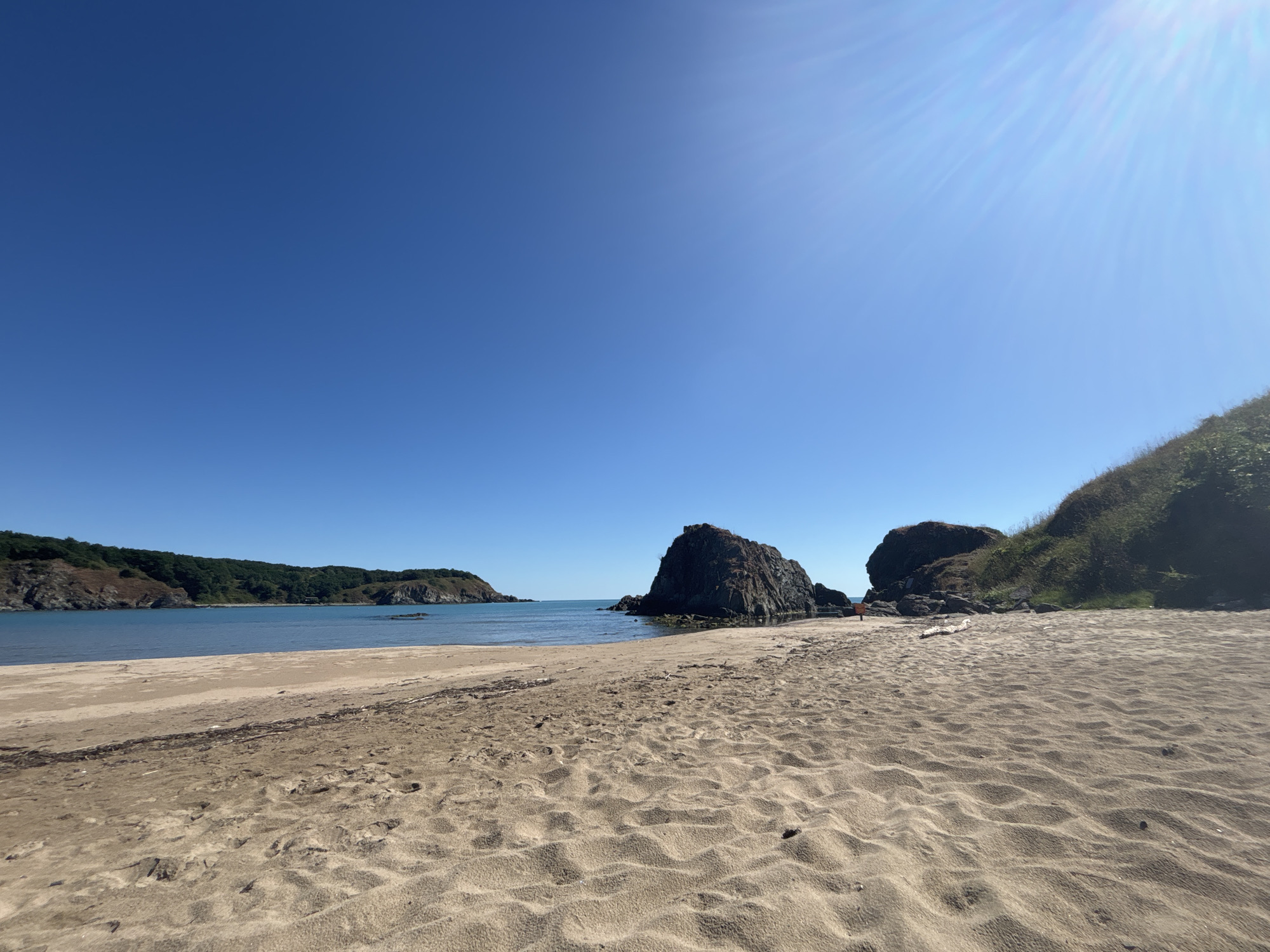 The image depicts a sandy beach in Rezovo, Bulgaria. The foreground is dominated by textured sand with footprints. In the mid-ground, the calm blue sea meets the shore, with several large dark rocks protruding from the water. Further in the distance, a green, tree-covered coastline forms a bay. The sky is a clear, bright blue with a sun flare visible in the upper right quadrant. The overall scene is a serene coastal landscape.