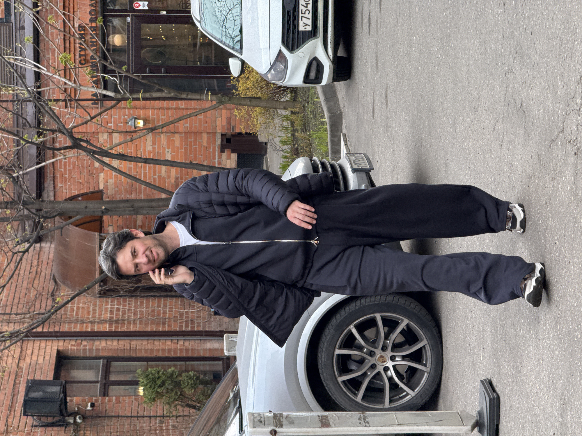 A man is standing on the hood of a silver Porsche Cayenne, talking on a cell phone. He is wearing a navy blue tracksuit and white and black sneakers. Behind him is a brick building with a sign in Cyrillic, likely indicating a studio or business name. A white Hyundai Solaris is parked nearby. The location appears to be in an urban setting in Moscow, Russia, based on the license plates of the vehicles.