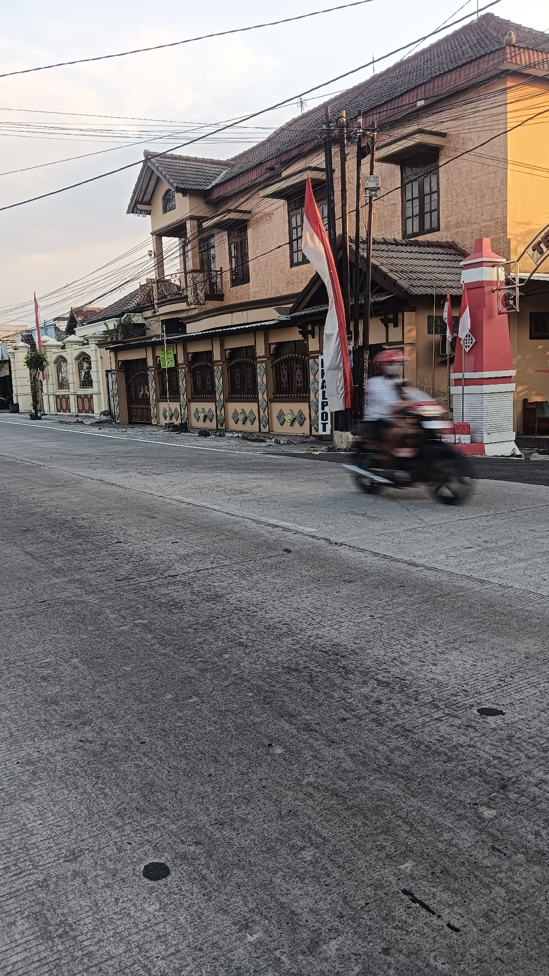 The image depicts a street scene in Sragen, Indonesia, with a focus on a building and a motorcycle. The building, a two-story structure with beige walls and a dark tiled roof, is located on the right side of the street. It features multiple windows with dark frames and a decorative balcony on the second floor. Two Indonesian flags are prominently displayed, one a large vertical flag on the left side of the building and a smaller one on a red and white pillar to the right. The street itself is paved with concrete and shows signs of wear, with visible lines and some darker patches. A person on a dark-colored motorcycle is captured in motion blur, traveling on the street from left to right. The sky above is overcast, with subtle hints of sunlight. The overall impression is of a typical urban street in the region, with elements of local culture and everyday activity.