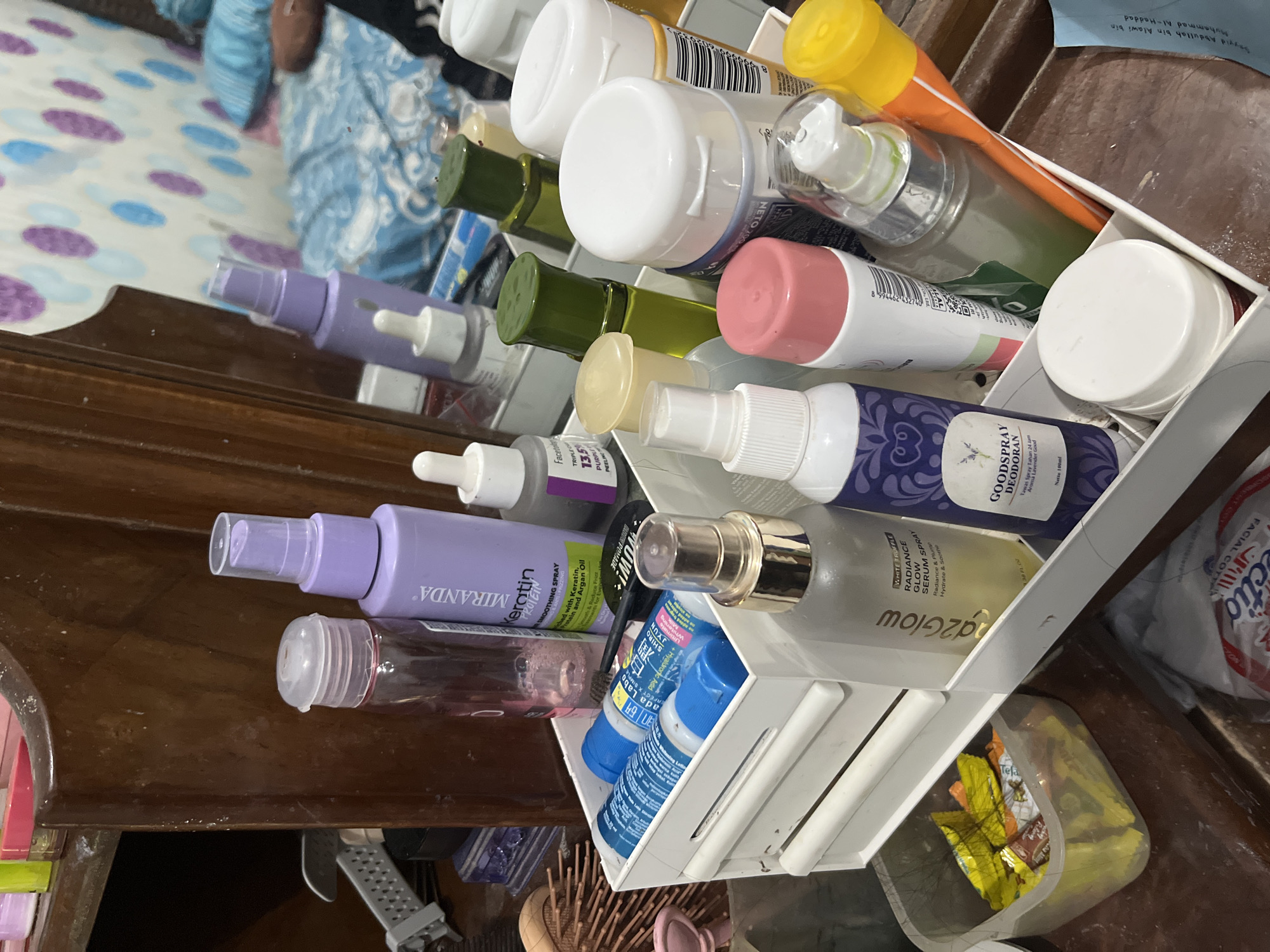 The image depicts a close-up view of a wooden dresser surface, likely in Glagah Kulon, Indonesia, cluttered with various personal care and cosmetic products. A white organizer tray holds several items, including two blue-capped bottles, a gold-capped serum spray, a purple-labeled deodorant spray, and a white jar with a lid. To the right of the tray, a green bottle, a white bottle with a black cap, and a yellow-capped orange-handled object are visible. In the background, a mirror reflects a wall with a patterned design of purple and blue circles. A pink-topped spray bottle and a clear bottle with pink liquid are also present on the dresser. A small clear plastic container filled with individually wrapped packets is situated to the left of the organizer tray. The overall scene suggests a personal vanity area.