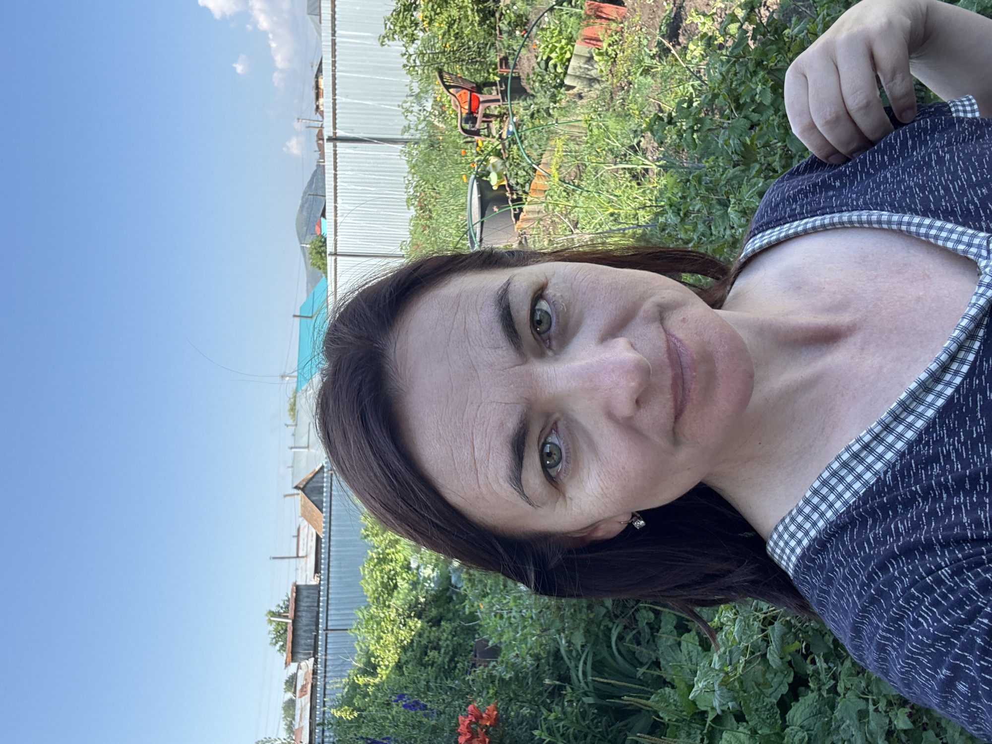 A medium-shot, outdoor, selfie-style photograph shows a middle-aged woman with dark brown hair, green eyes, and fair skin. She is looking directly at the camera and smiling slightly. She is wearing a dark blue shirt with a white checkered collar. The background is a garden with lush green plants, a metal fence, and some buildings in the distance. The sky is clear and blue. The scene is likely located in the city of Urazovo, Russia, as indicated by the presence of dacha-style buildings and a garden common in rural Russian settings.