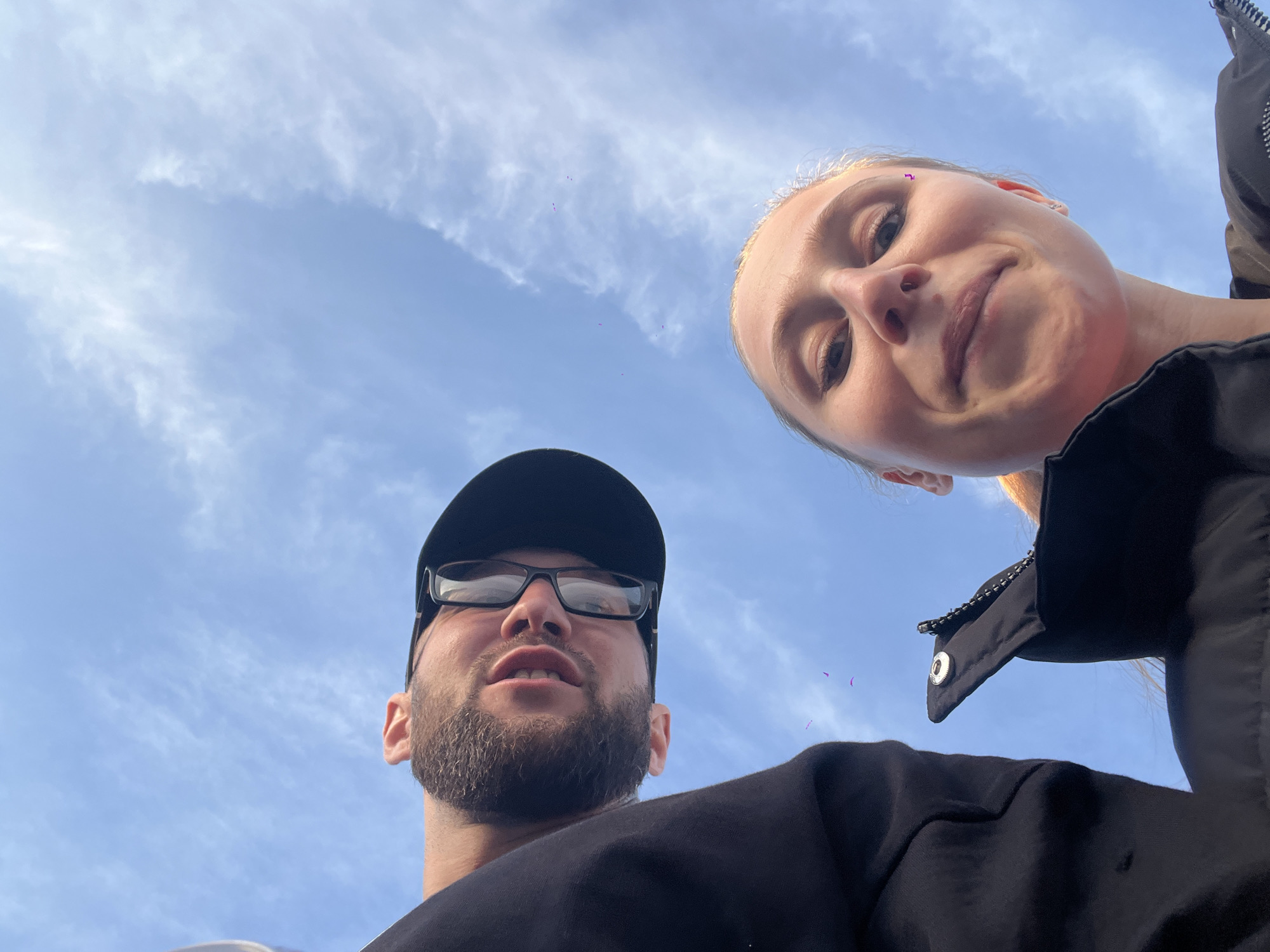 The image shows two people from a low angle perspective. A man with a beard, glasses, and a black baseball cap is on the left. A woman with blonde hair is on the right. Both are wearing black clothing. Above them, a blue sky is visible with scattered clouds. The location is not identifiable as Budva, Montenegro.